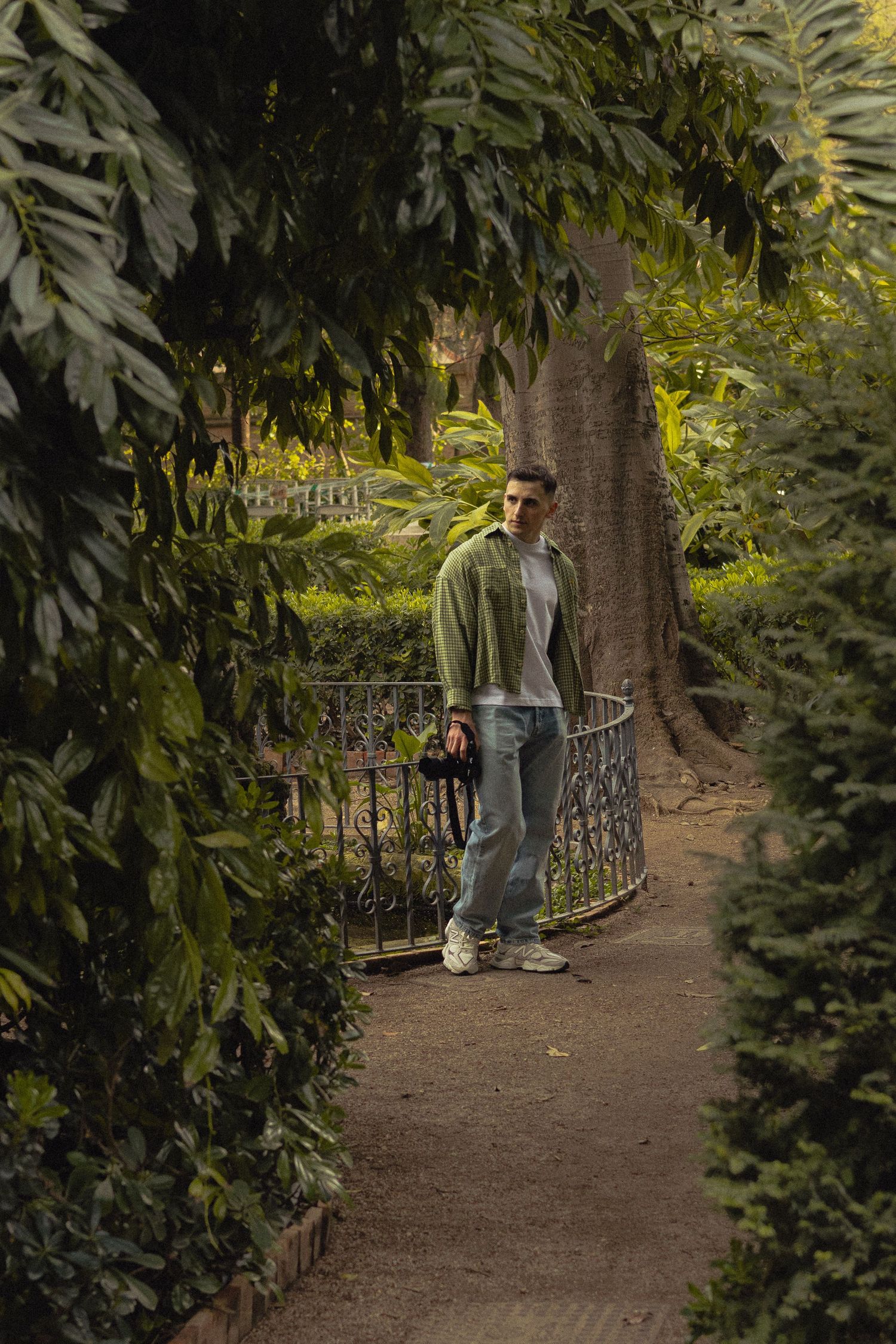 Man standing on a garden path surrounded by lush tropical greenery and iron fence.