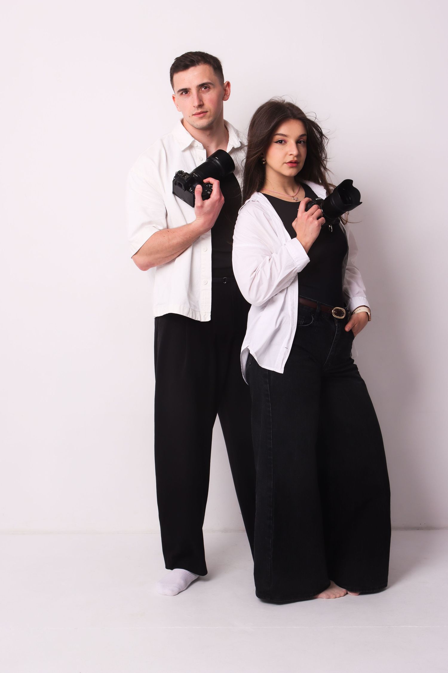 A stylish couple in black and white outfits hold cameras while posing against a white wall.