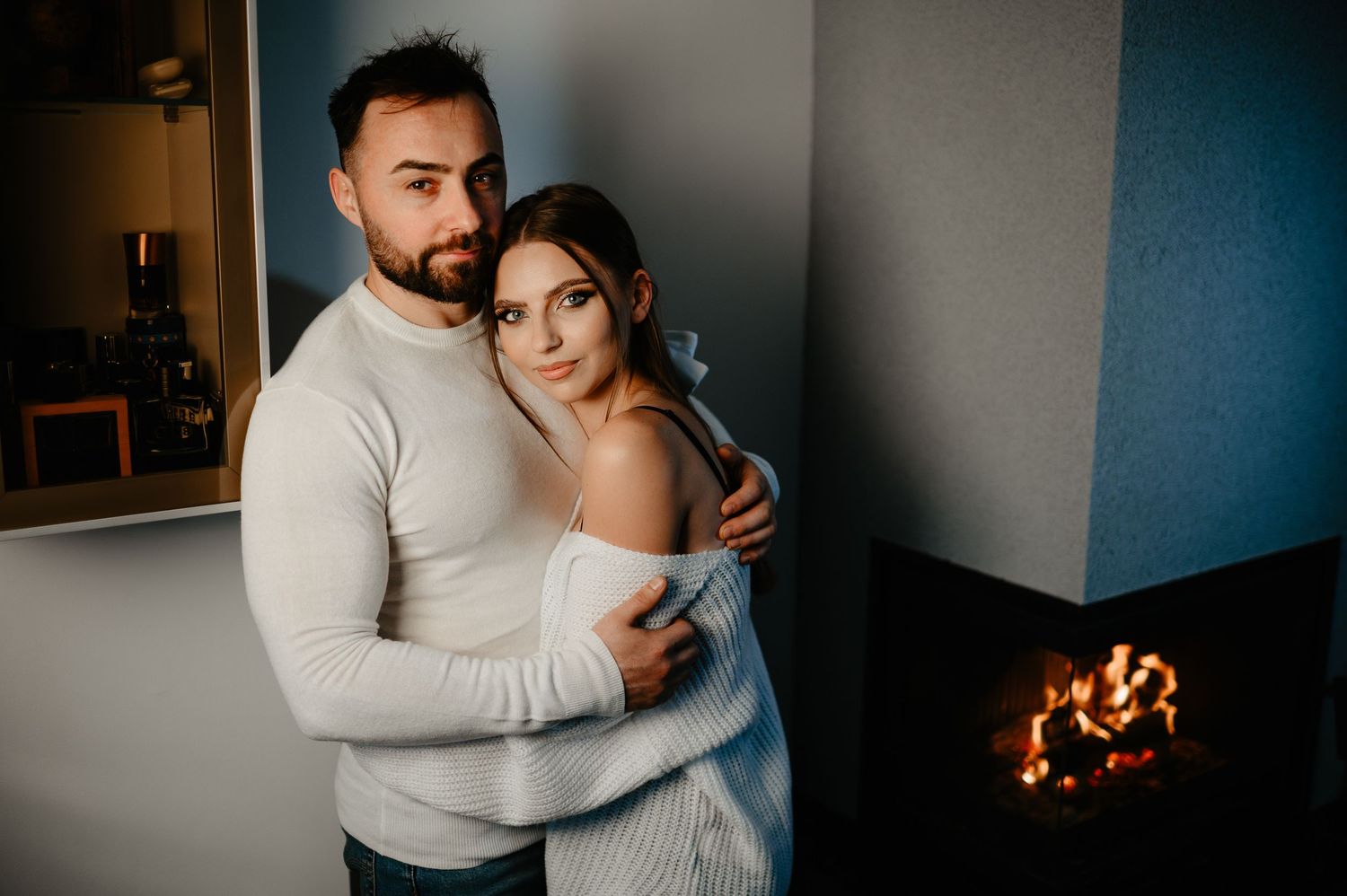 Couple in white sweaters embrace lovingly beside a modern fireplace in a stylish interior.