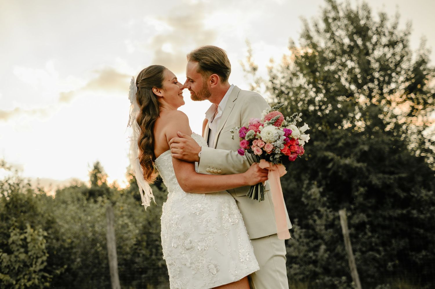 Bride and groom sharing a romantic kiss at sunset surrounded by trees holding colorful bouquet.