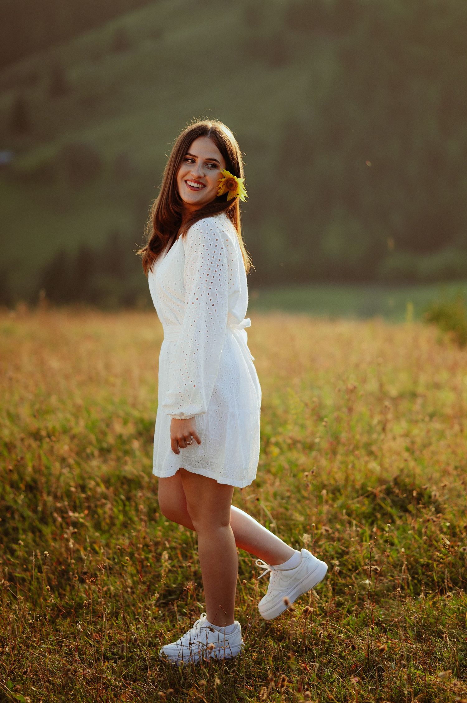 Young woman in white dress smiling over shoulder in golden sunset mountain meadow.