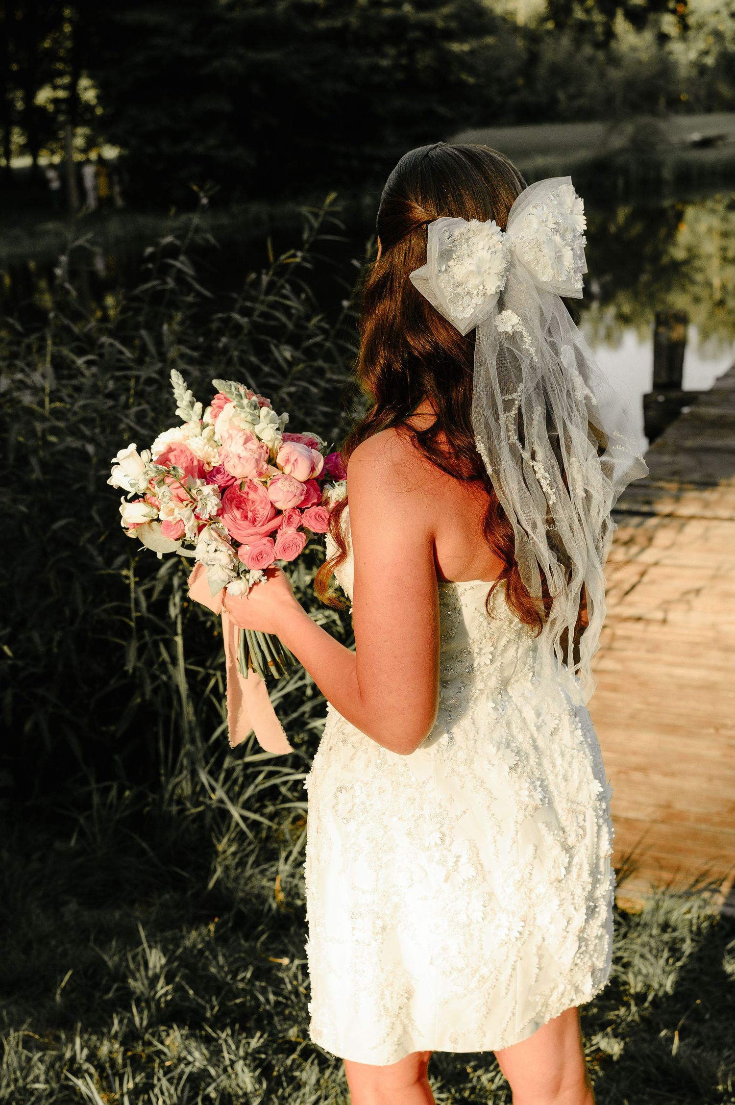 Bride in short white lace dress with large bow veil holds pink bouquet near a sunlit lakeside dock.