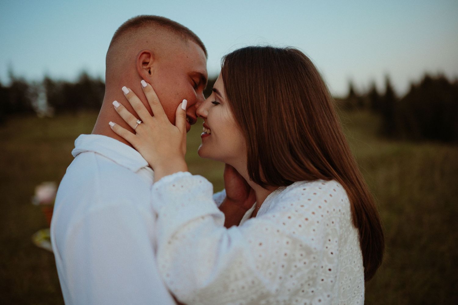 Romantic couple almost kissing outdoors at dusk, woman with engagement ring embracing mans face in green meadow.