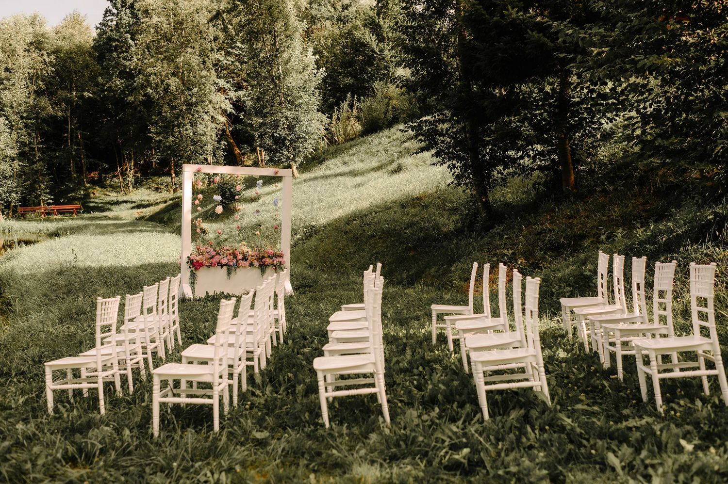 Elegant outdoor wedding ceremony with white chairs and floral arch on a green hillside.