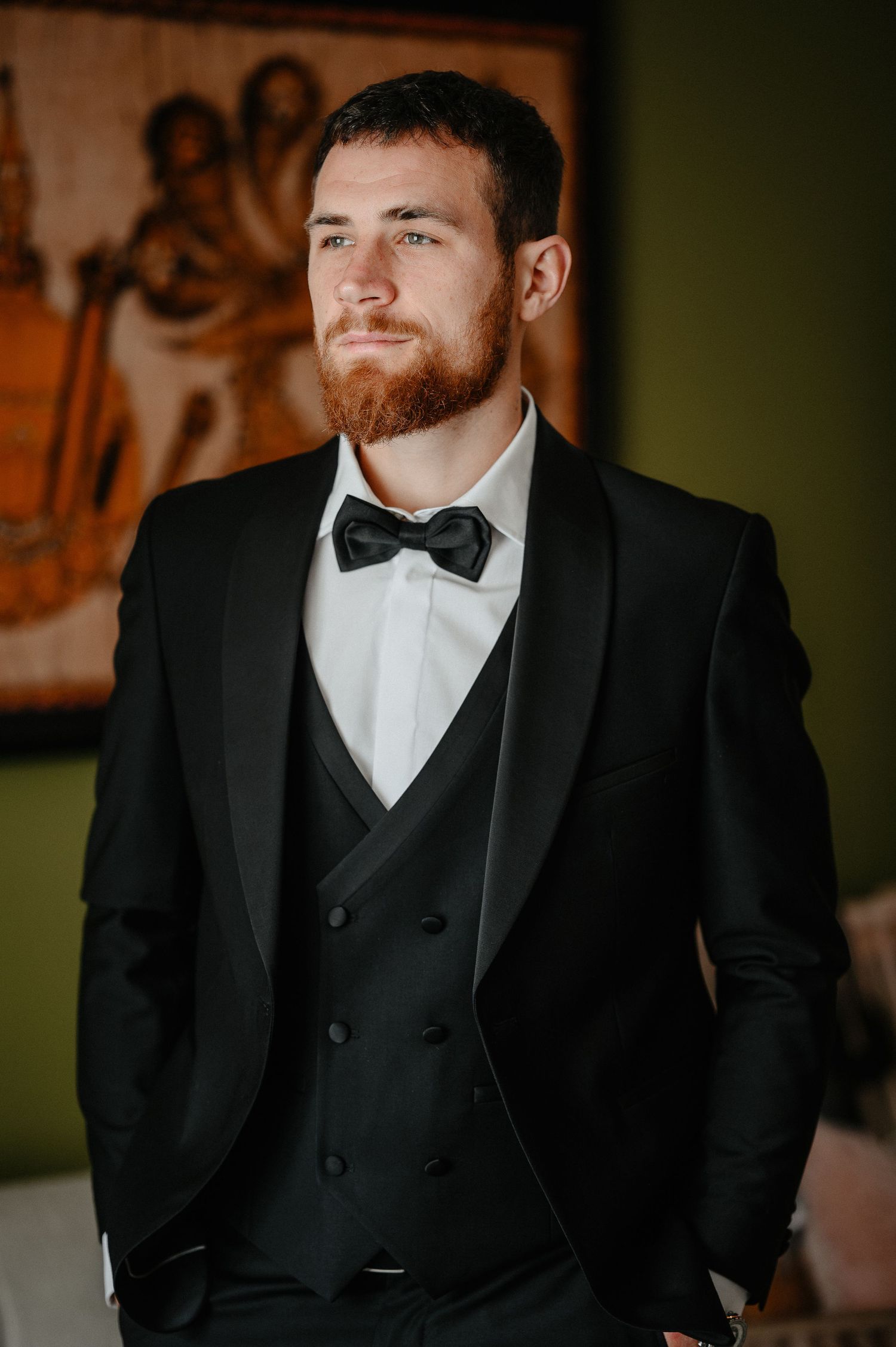 Handsome bearded groom in a black tuxedo with bow tie posing confidently in a hotel room.