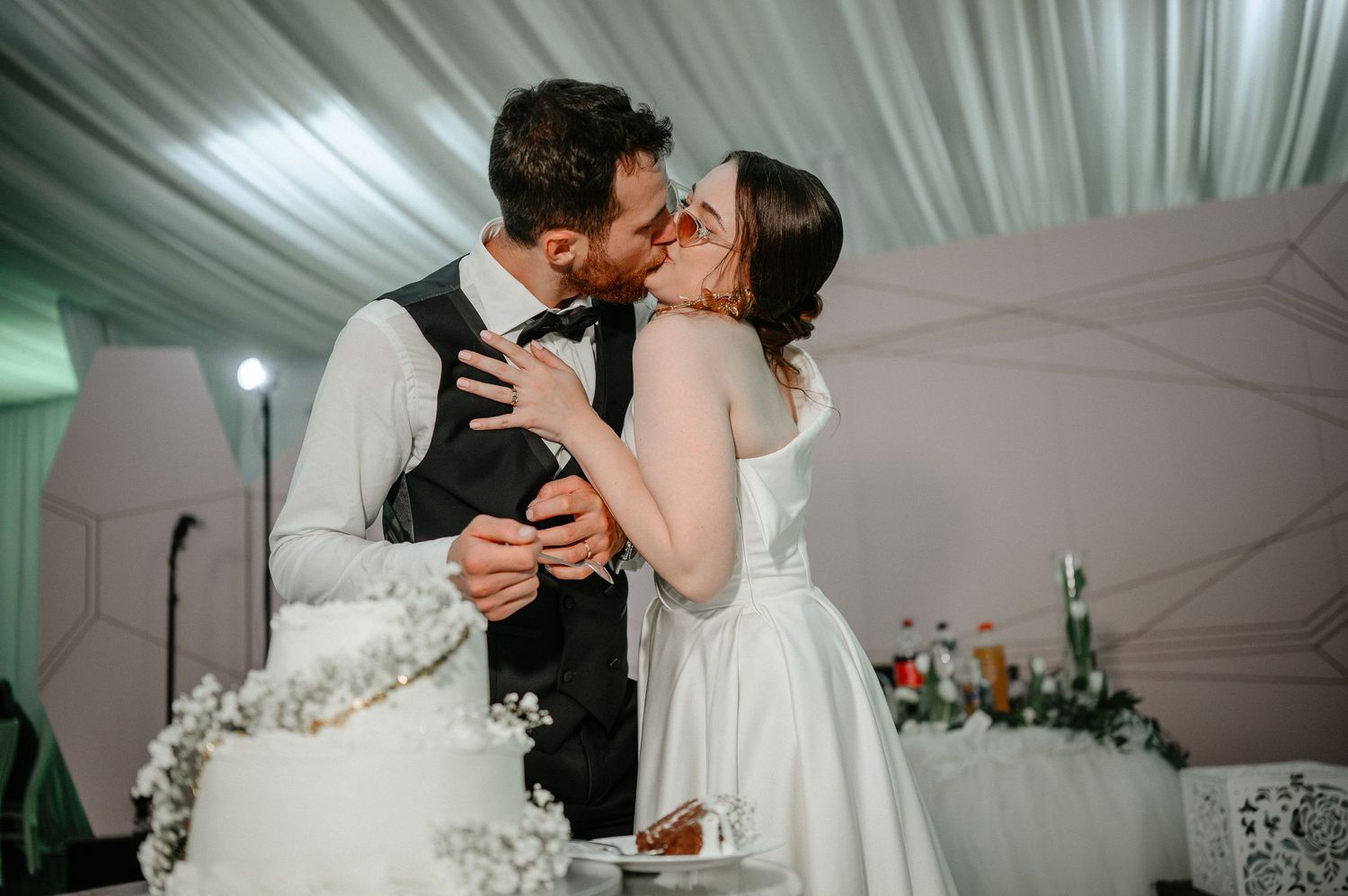 Bride and groom sharing a romantic kiss beside their white wedding cake at indoor reception.