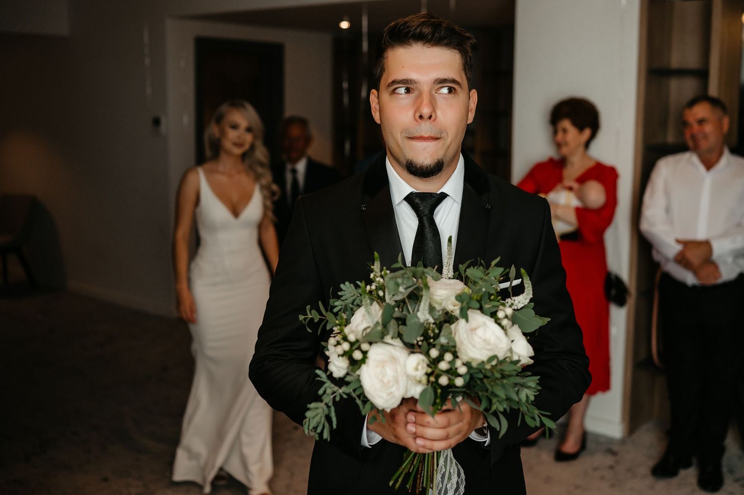 Groom in black suit holding elegant white rose and greenery bridal bouquet before wedding ceremony.