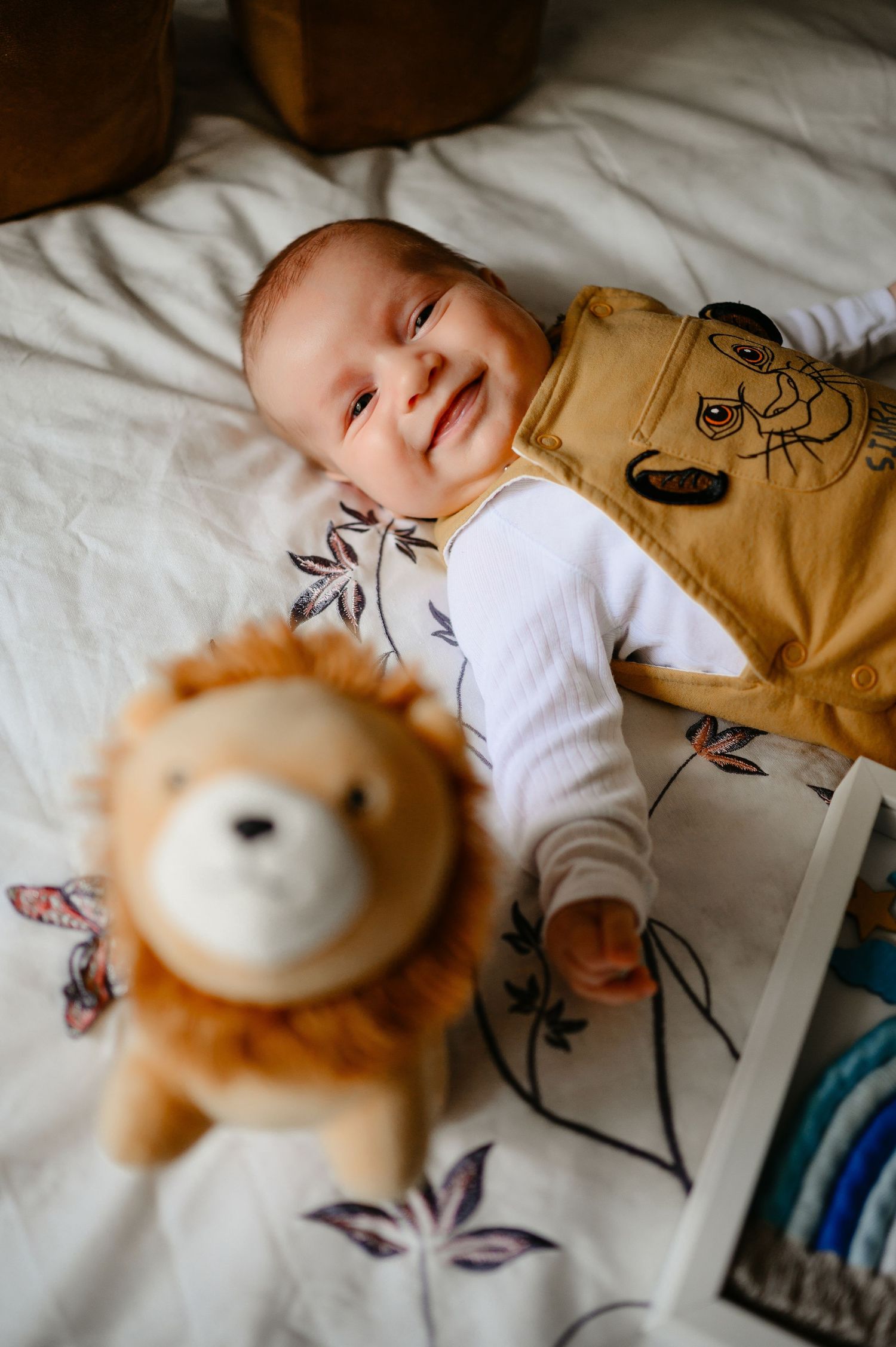 Smiling baby lying on white floral blanket next to a plush lion stuffed animal toy.