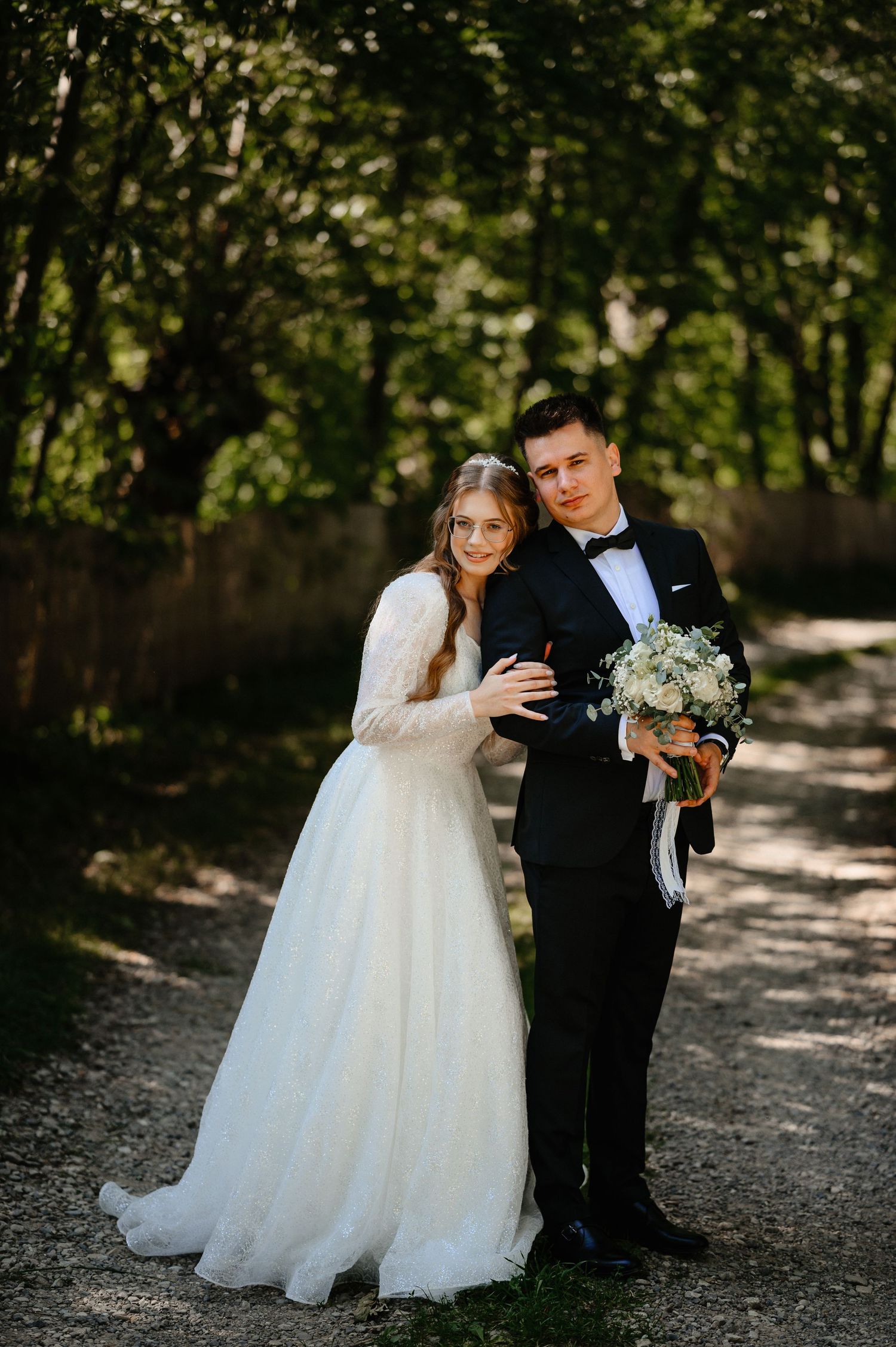 Bride in white lace gown and groom in tuxedo posing together on garden path.