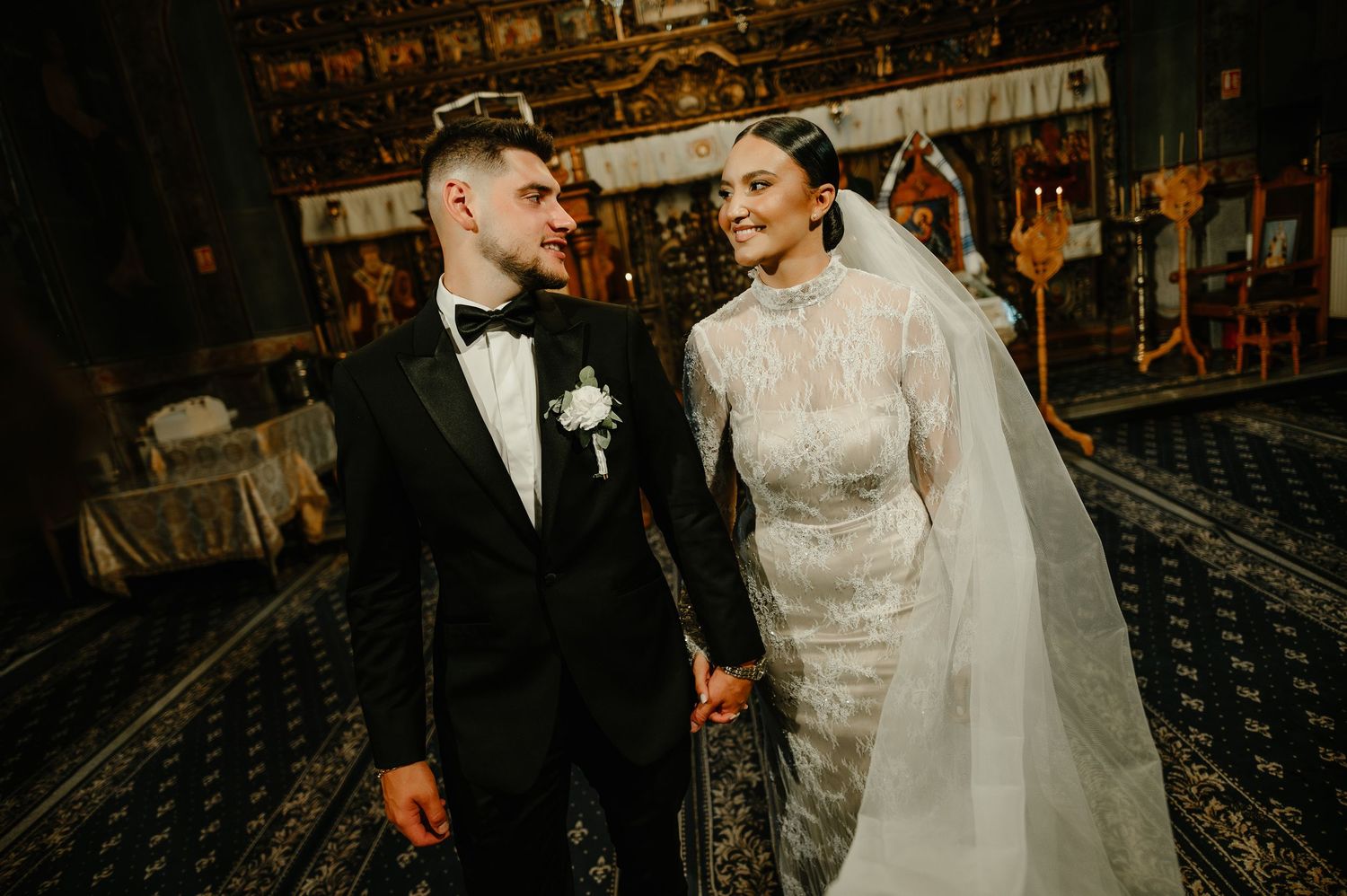 Bride and groom gazing at each other lovingly while holding hands inside a richly decorated church.