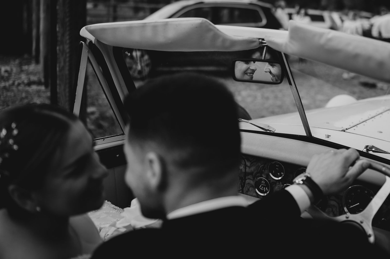 Black and white close-up of newlyweds kissing beside a classic vintage white wedding car.
