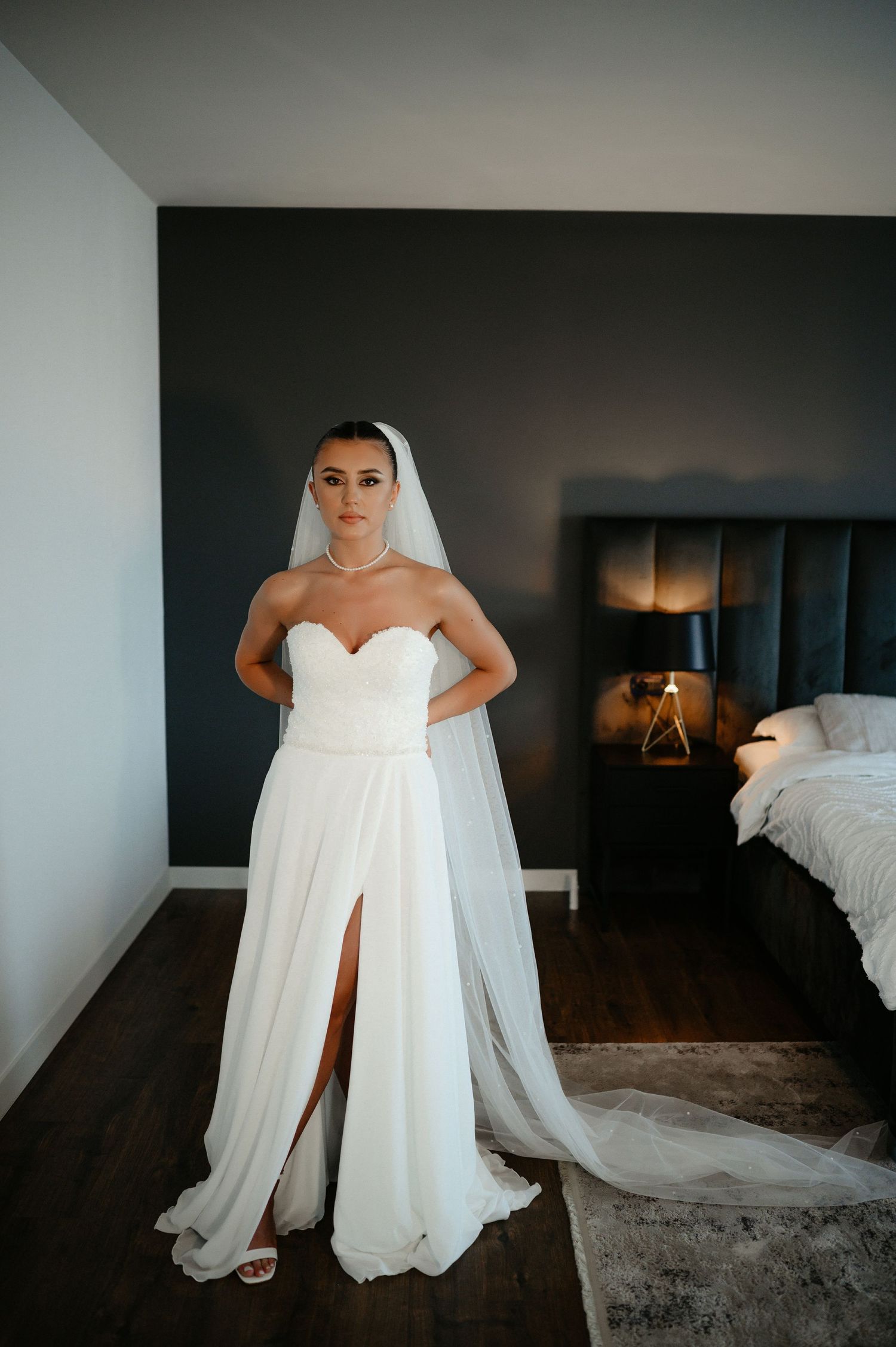 Elegant bride in strapless white gown with veil and slit skirt in bedroom.