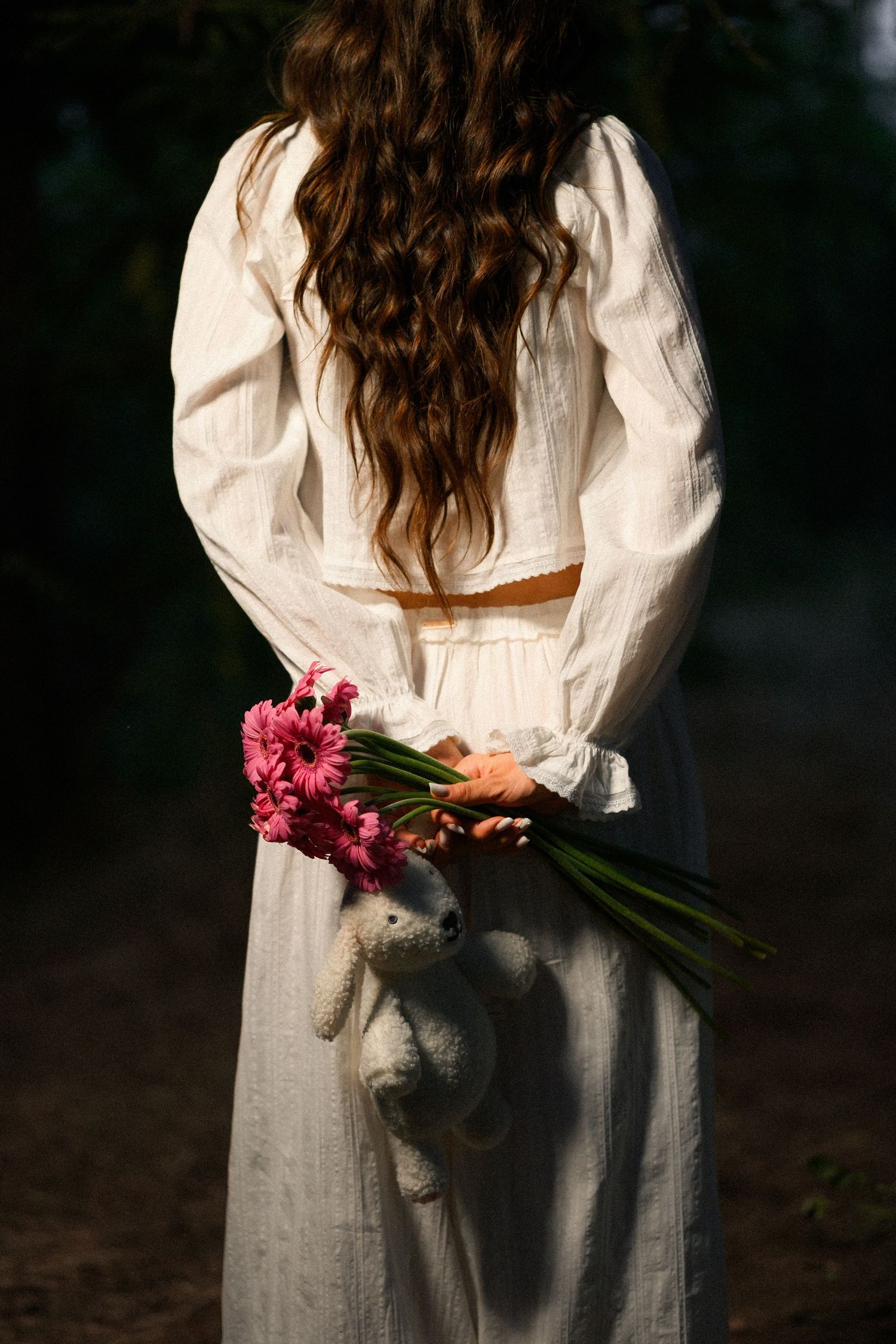 Girl in white dress holding pink gerbera flowers and a stuffed bear behind her back, standing outdoors.