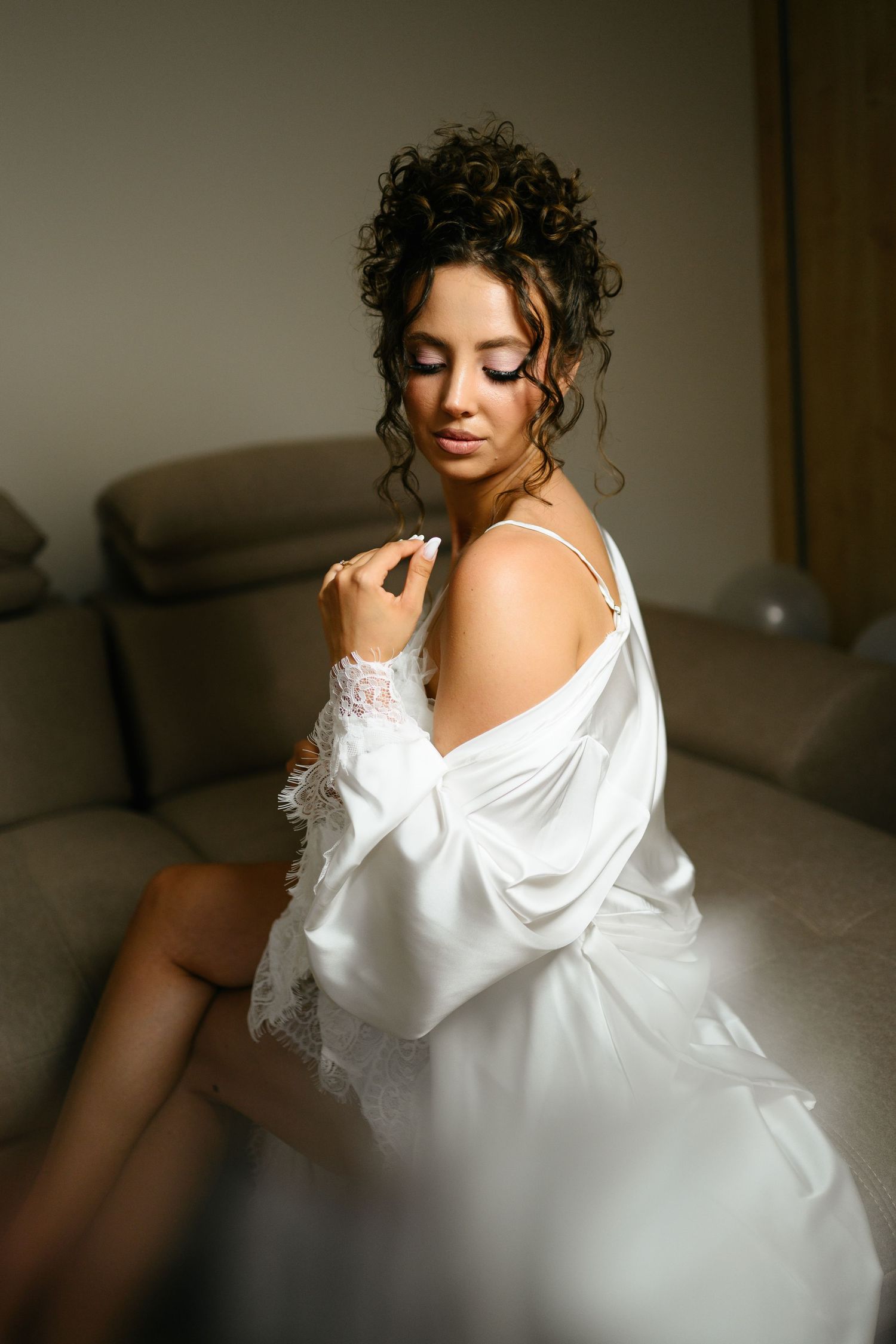 Elegant bride in white satin robe with lace trim, sitting on sofa with updo hairstyle and glamorous makeup.