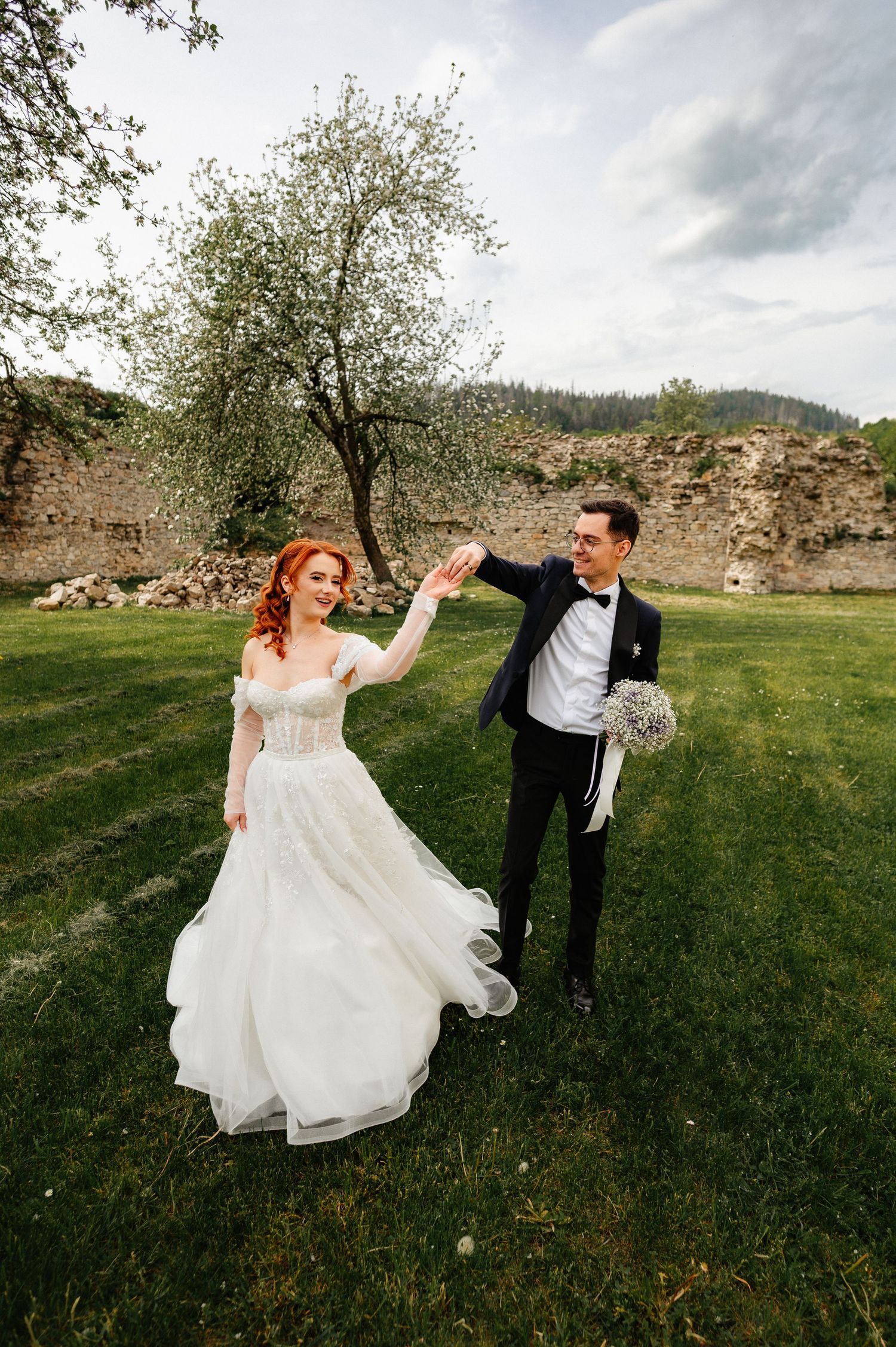 A bride in a white gown and groom in a tuxedo dance on green grass near ancient stone ruins and a blooming tree.