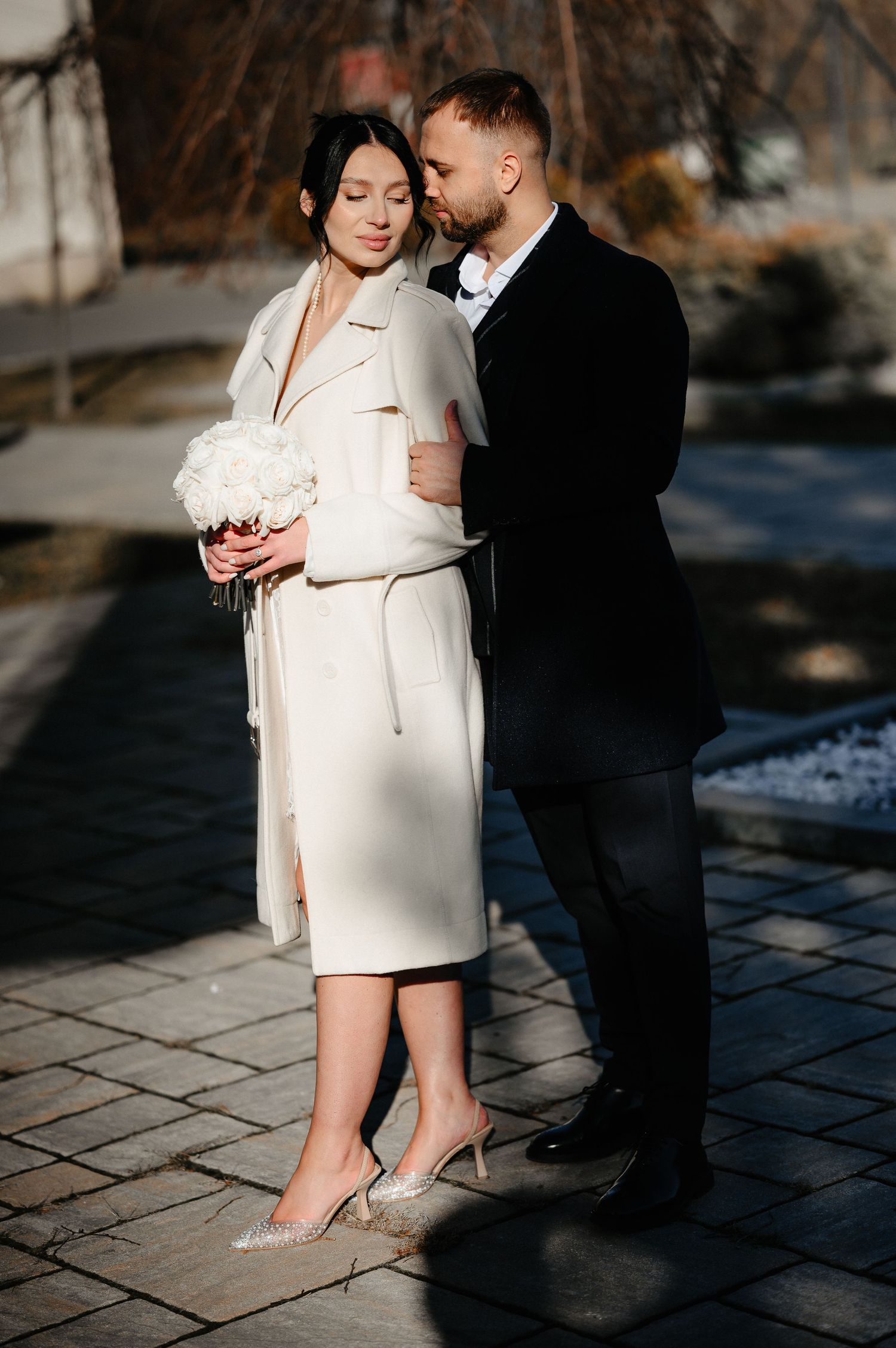 Romantic couple embracing outdoors, bride in white coat holding white bouquet, groom in black suit.