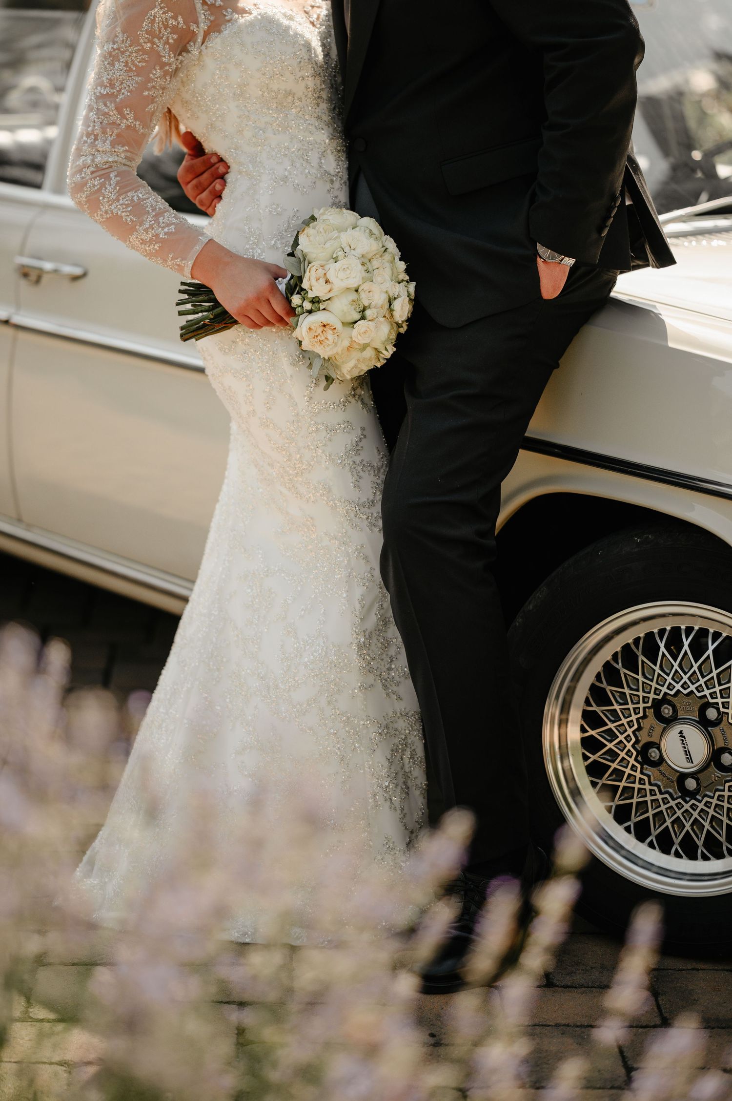 Bride in fitted lace wedding dress holding white bouquet standing beside groom next to classic car.
