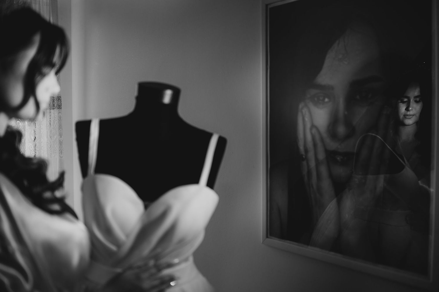 Black and white photo of woman near dress form mannequin with wedding gown, reflected in framed artwork.