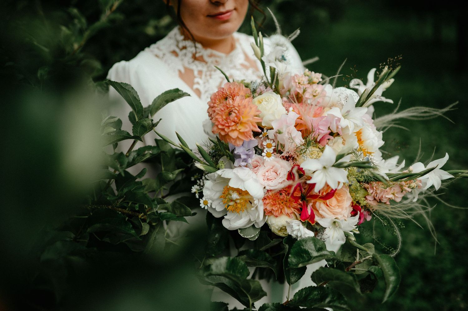 Bride holding a lush wedding bouquet with pink dahlias, white peonies, and greenery outdoors.
