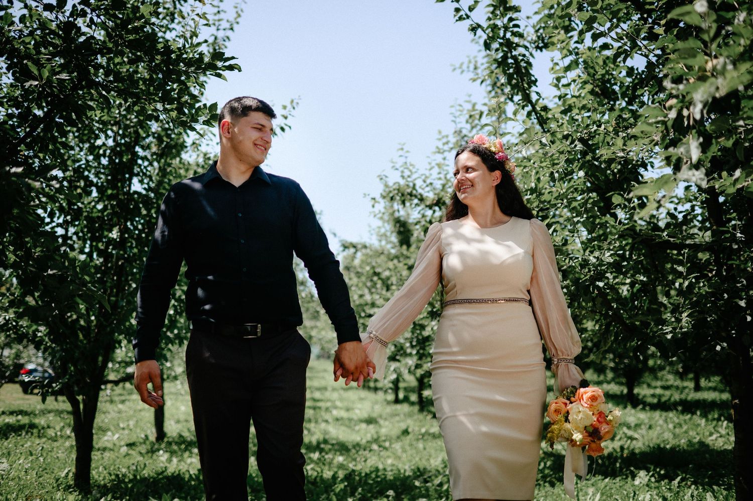 Bride and groom holding hands walking through a sunny green orchard, bride holding a peach rose bouquet.