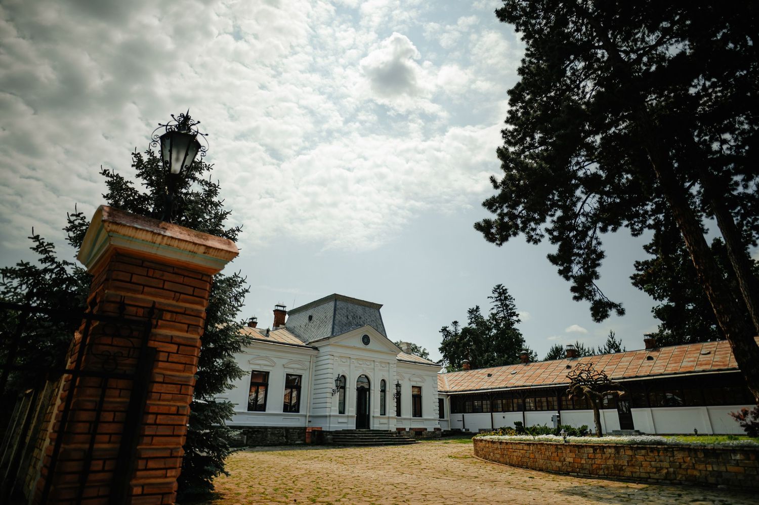 Historic white manor house with brick gate pillar and tall trees under cloudy sky.