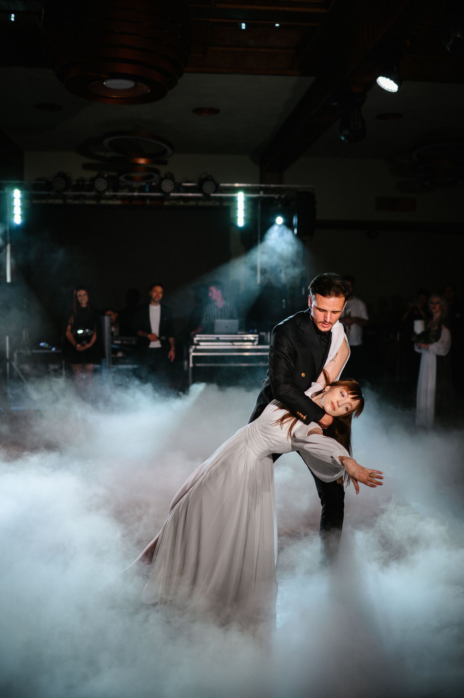Groom dips bride dramatically during first dance in a fog-filled wedding reception venue with stage lights.