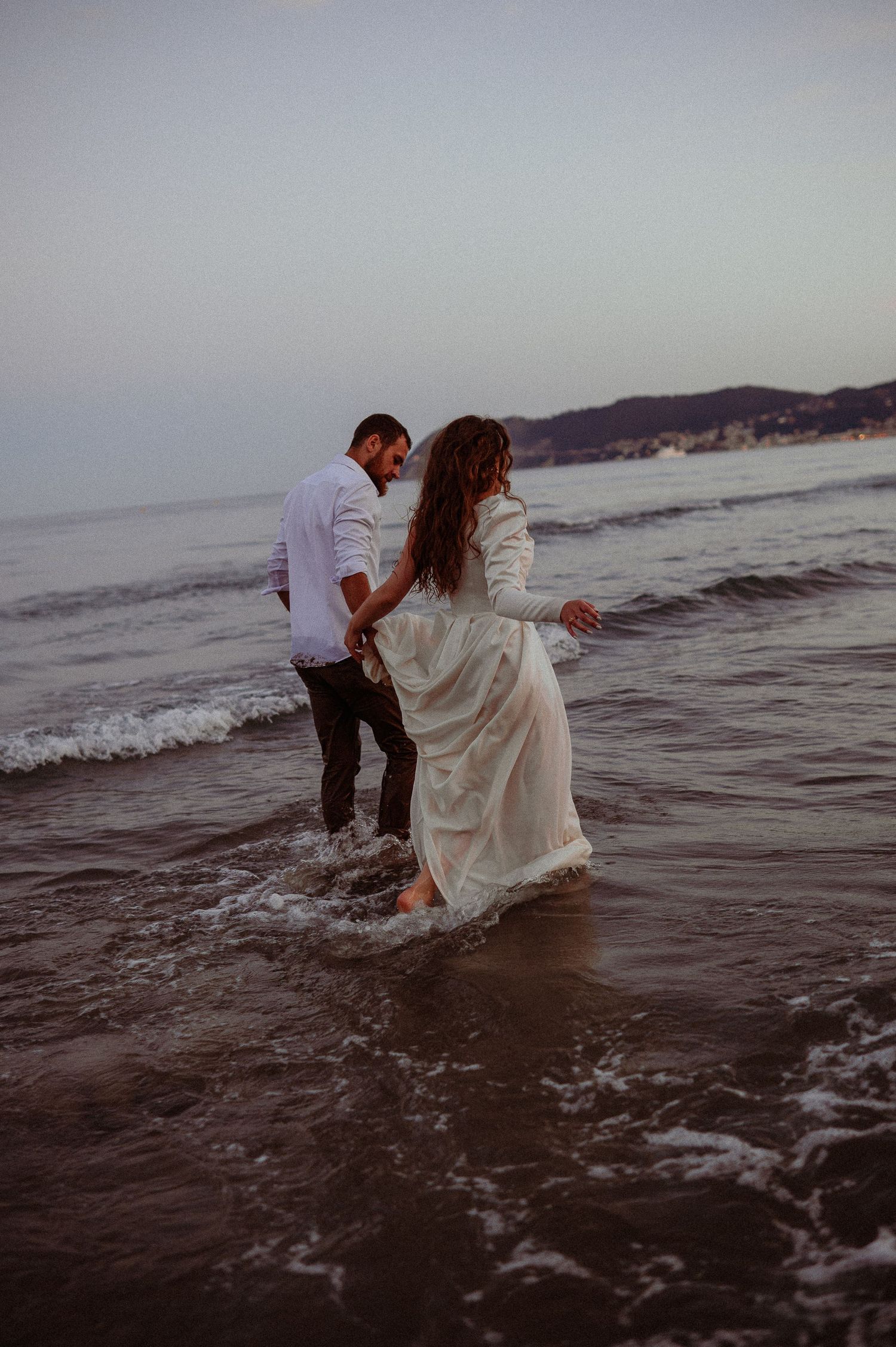 Couple holding hands and walking through shallow ocean waves on sandy beach at dusk in wedding attire.