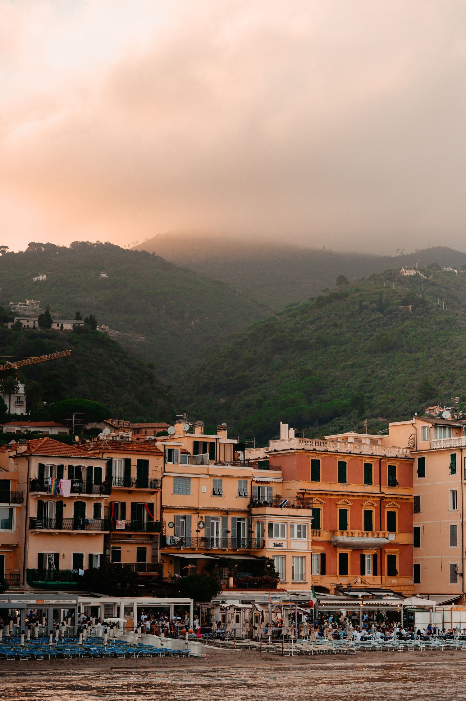 Warm sunset light illuminating pastel-colored Italian seaside buildings with beach umbrellas below green hills.