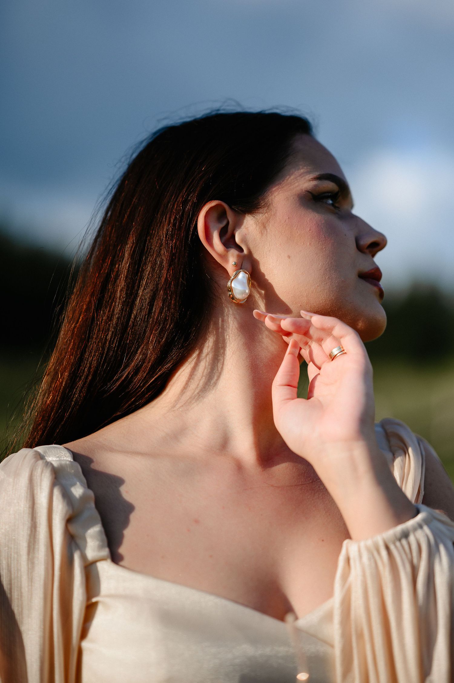 Portrait of brunette woman in cream dress touching her pearl earring against a cloudy sky.