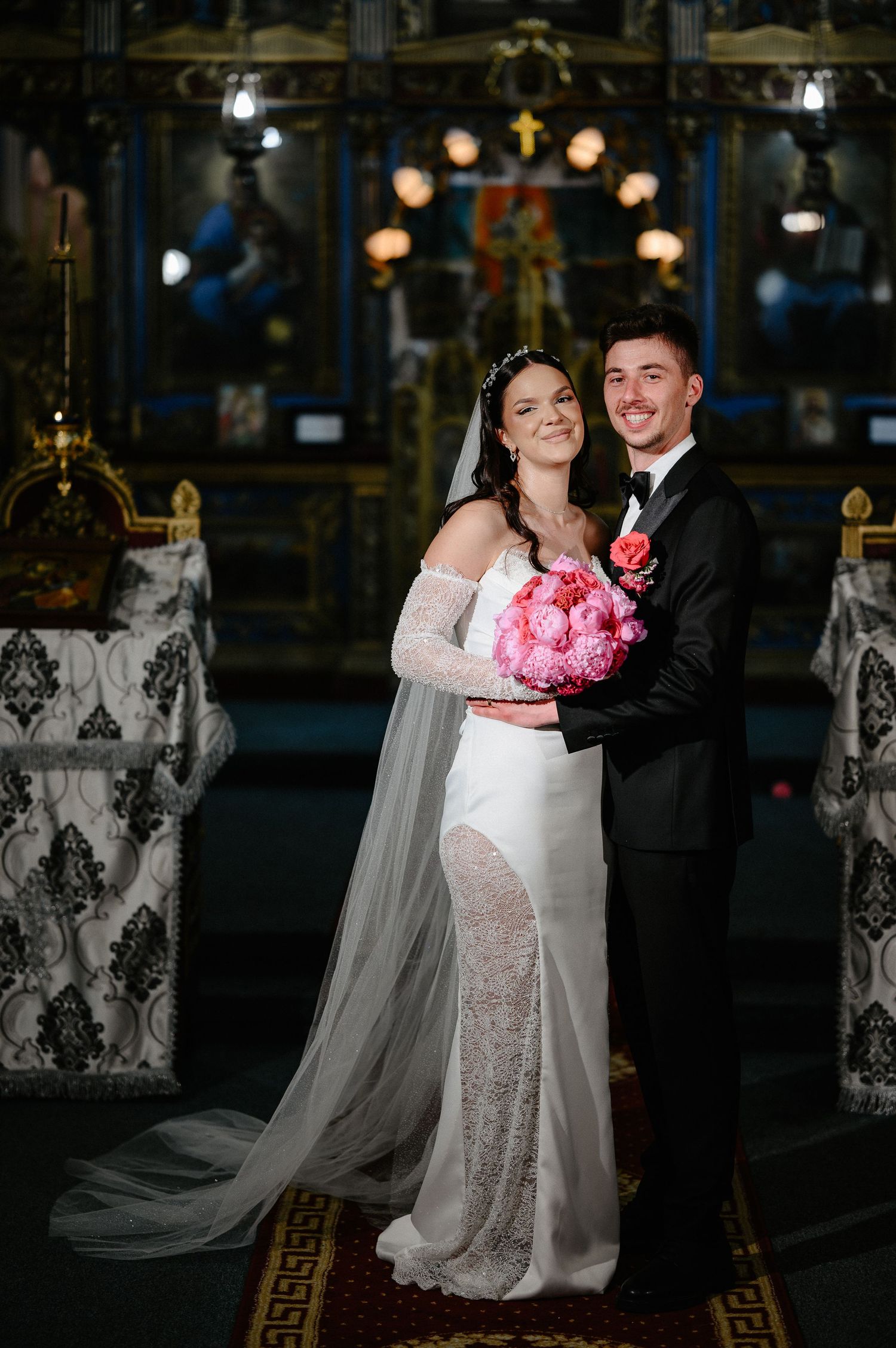 Bride in off-shoulder lace wedding gown holds pink bouquet with groom in black tuxedo in church.