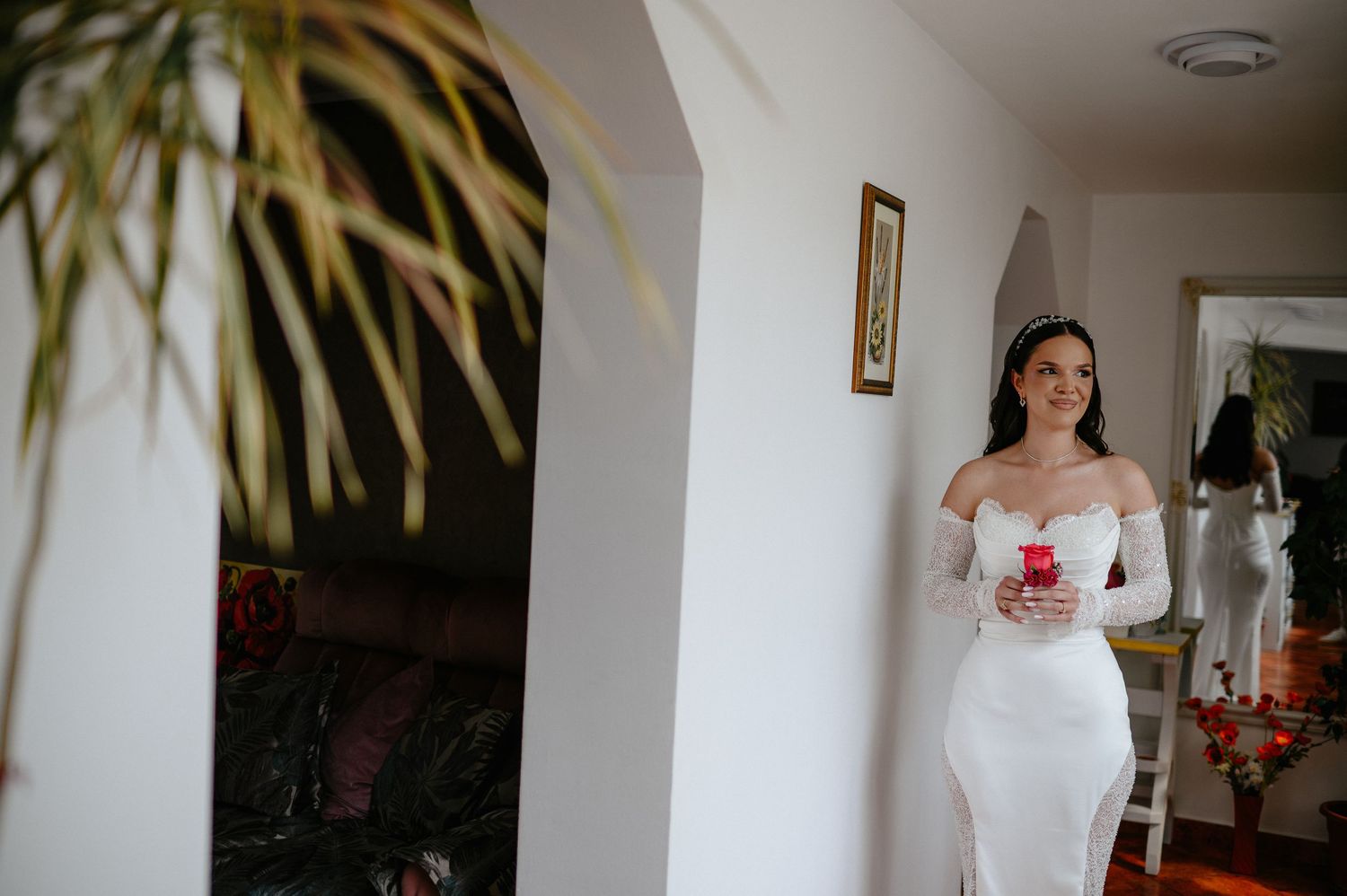 Bride in white off-shoulder gown holding red rose box in elegant home interior.