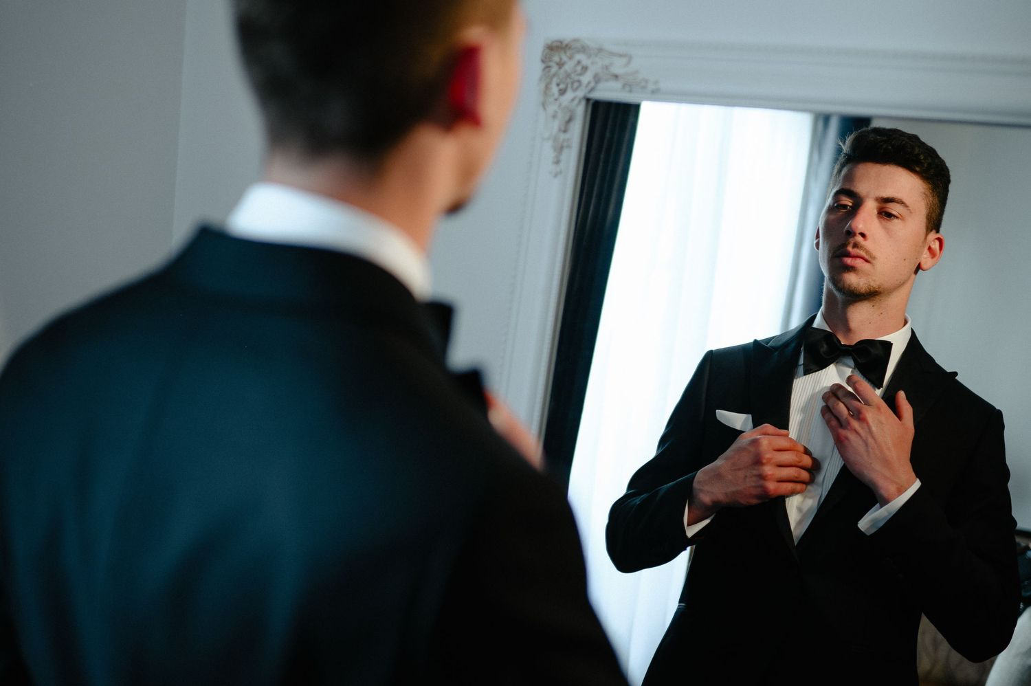 Groom adjusting bow tie while looking at his reflection in mirror before wedding.