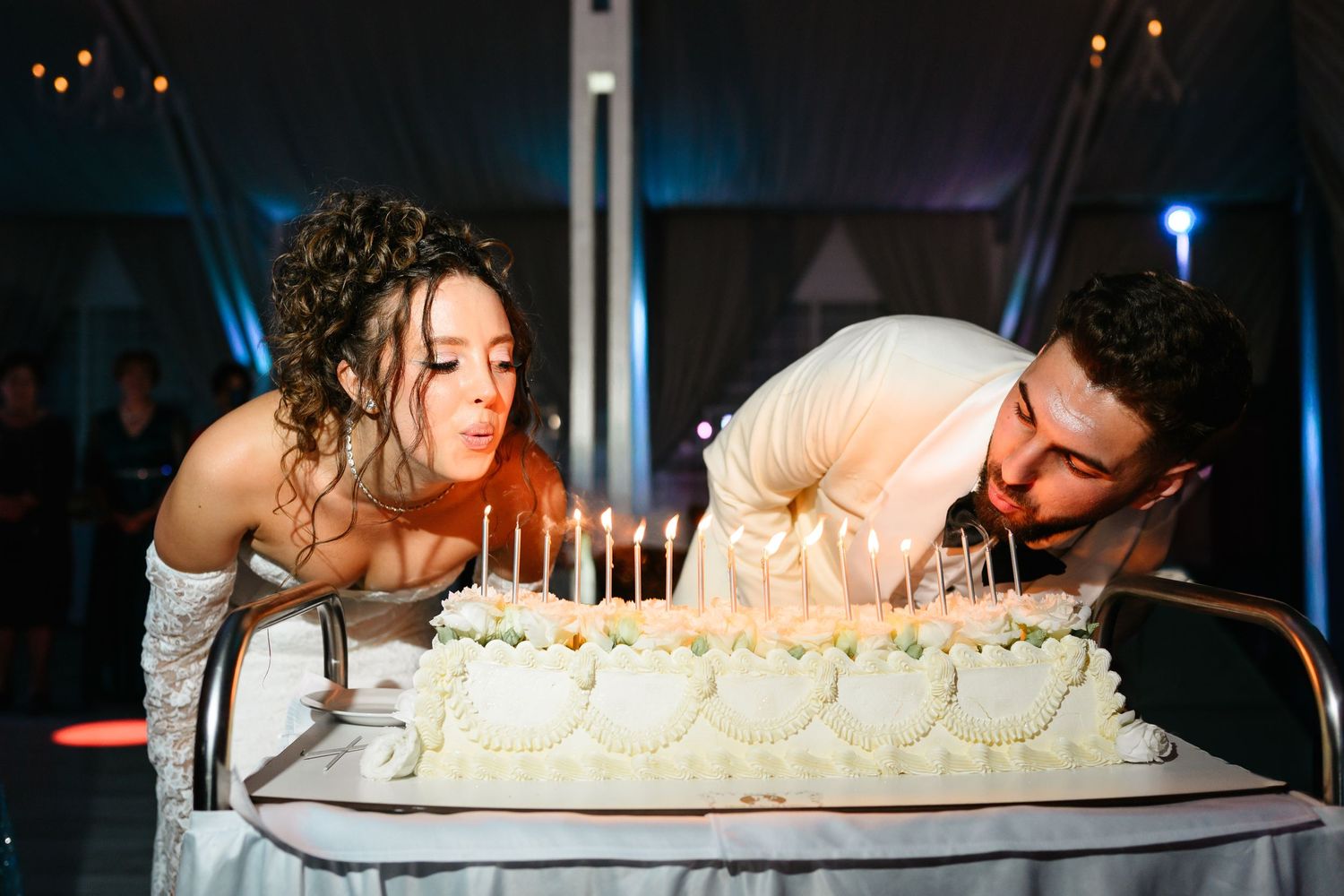 Bride and groom blowing out candles on a white wedding cake at their elegant evening reception.