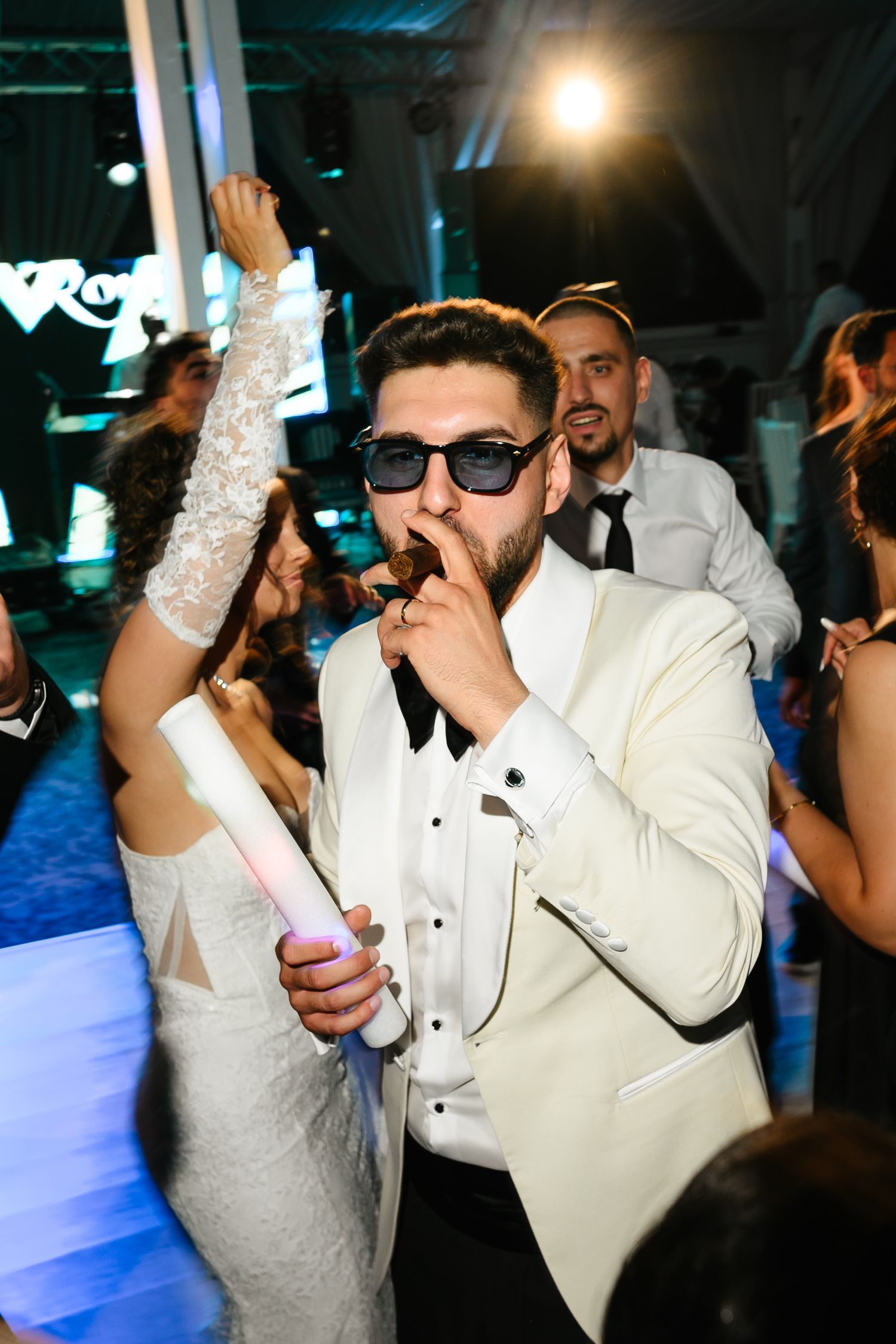 Groom in white tuxedo smoking cigar on wedding dance floor, wearing sunglasses, bride in lace dress beside him.