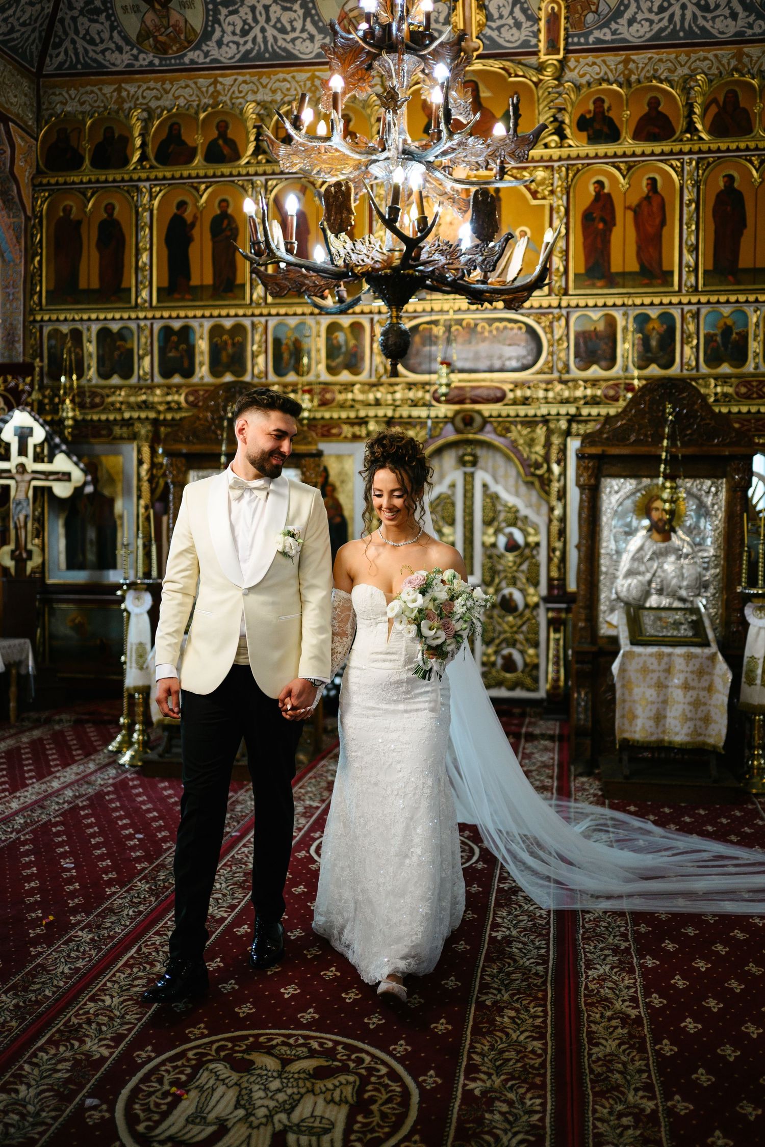 Bride and groom holding hands in an ornate Orthodox church, bride wearing lace gown with veil, groom in ivory tuxedo.