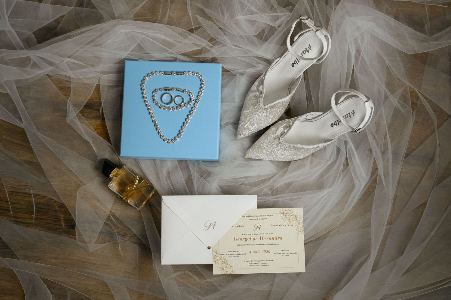 Elegant wedding flat lay with white heels, blue jewelry box, pearl necklace, perfume, and invitation on sheer veil.