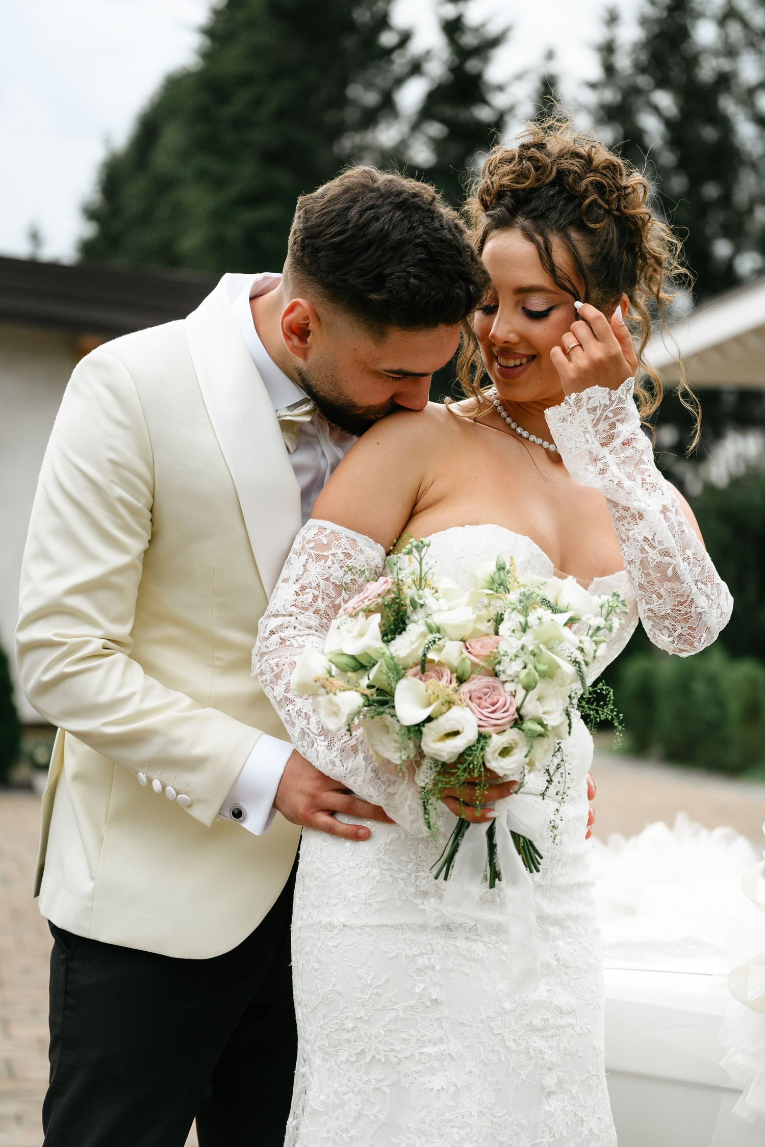 Bride and groom embrace outdoors, she holds white and pink bouquet, wearing lace gown, he in cream tuxedo jacket.