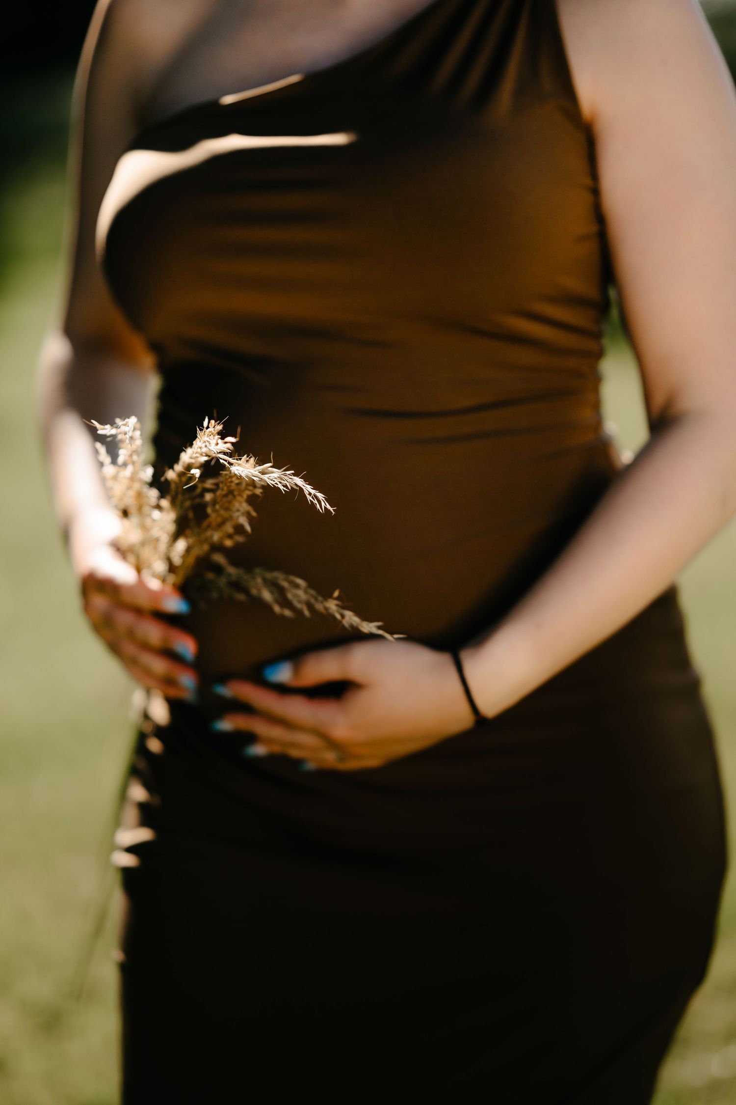 Close-up of pregnant woman in brown dress gently cradling baby bump while holding dried wildflowers outdoors.