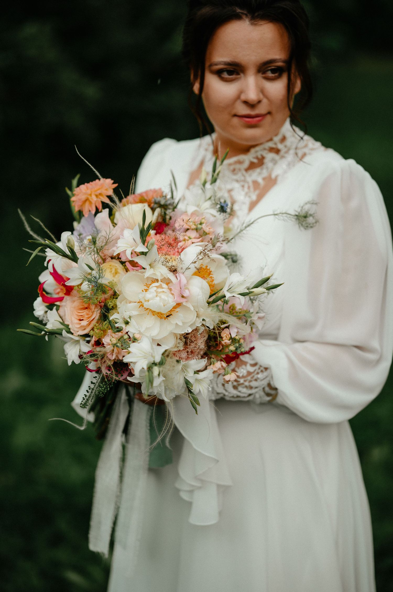 Bride in white lace dress holding colorful wedding bouquet outdoors.