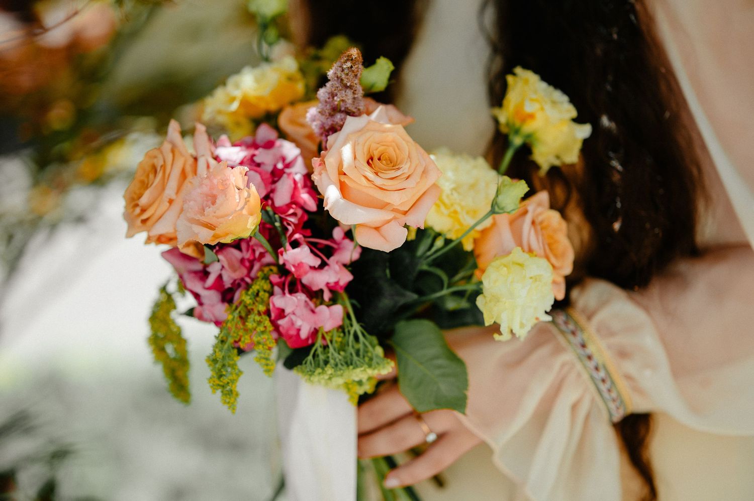 Bride holding a vibrant bouquet of peach roses, pink flowers, and greenery in a soft-focus outdoor setting.