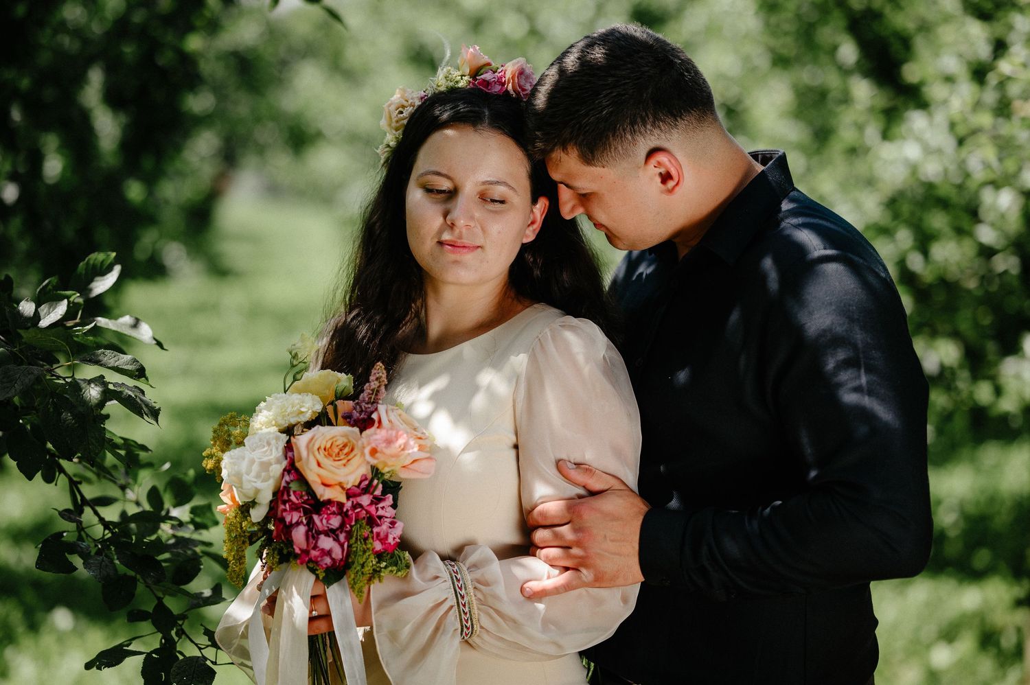 Bride in floral crown holds bouquet as groom in black jacket embraces her tenderly in lush green garden setting.