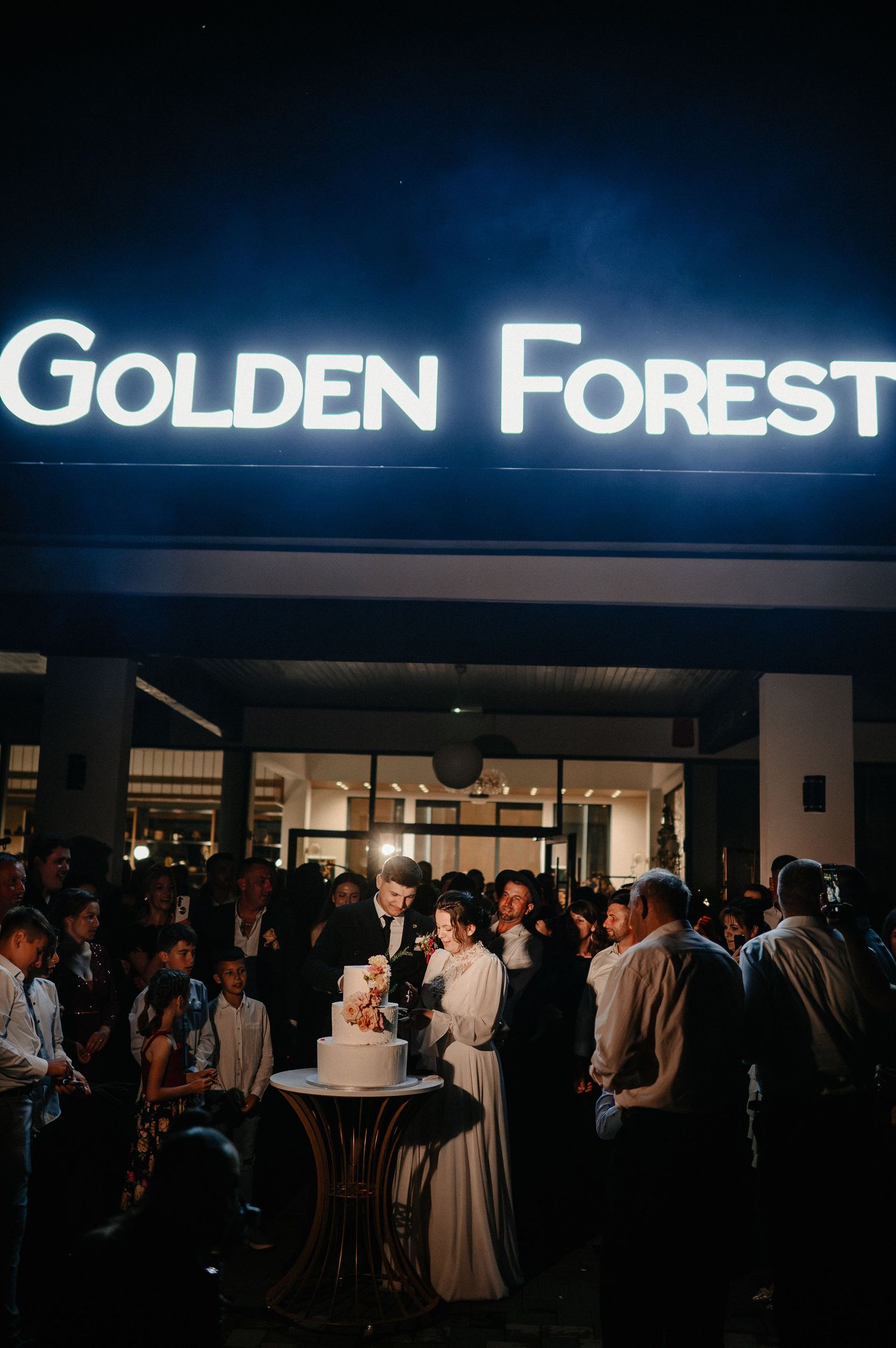 Couple cutting wedding cake outside illuminated Golden Forest venue at night.