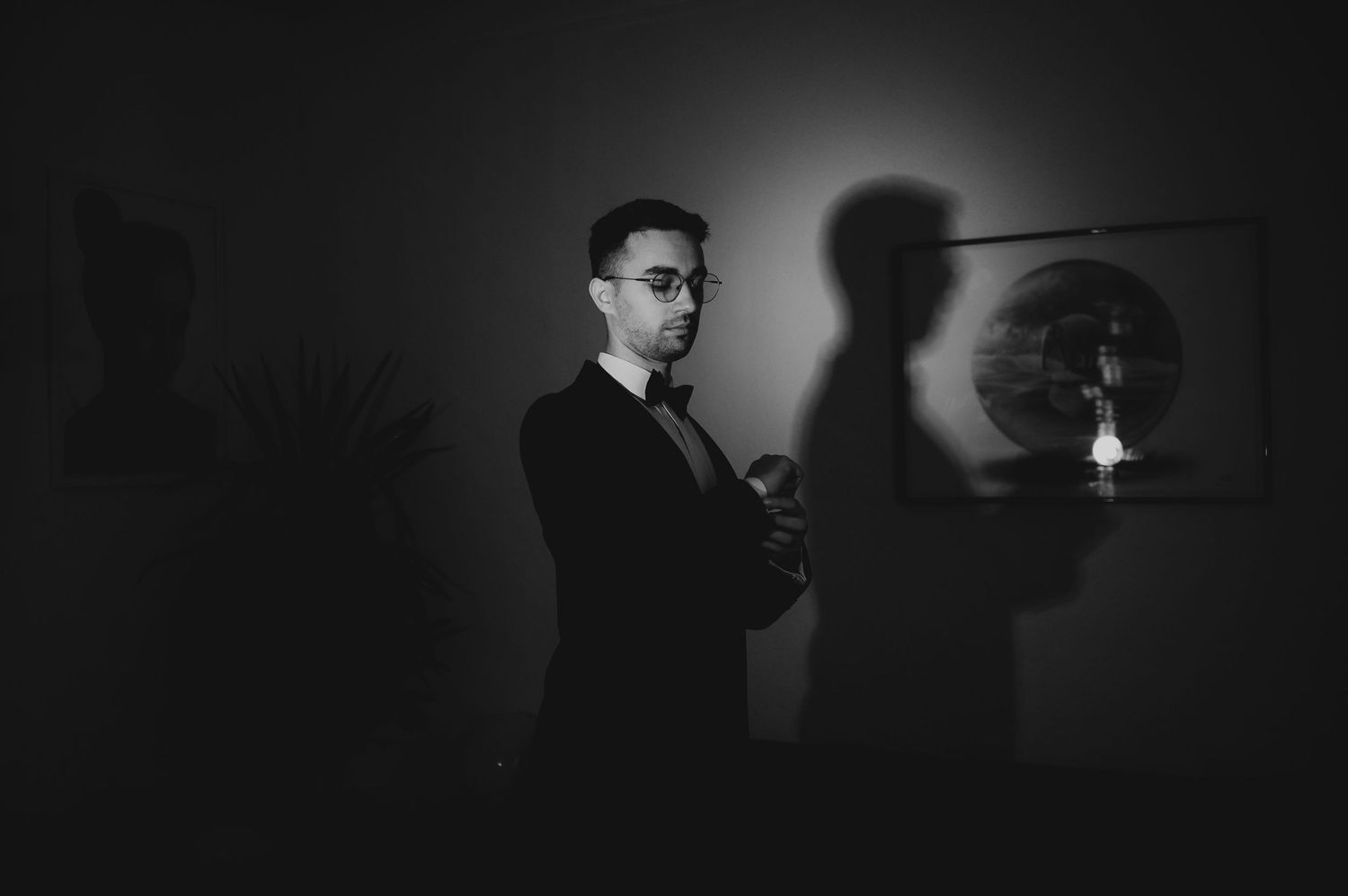Black and white portrait of a man in a suit with glasses, standing against a wall with dramatic shadow.