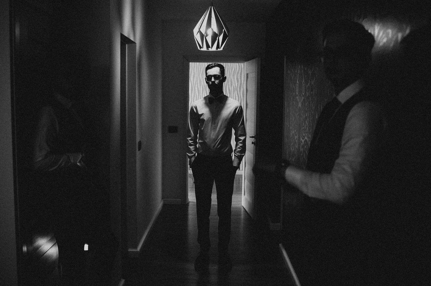 Black and white photo of a man standing in a dimly lit hallway with a pendant light above him.