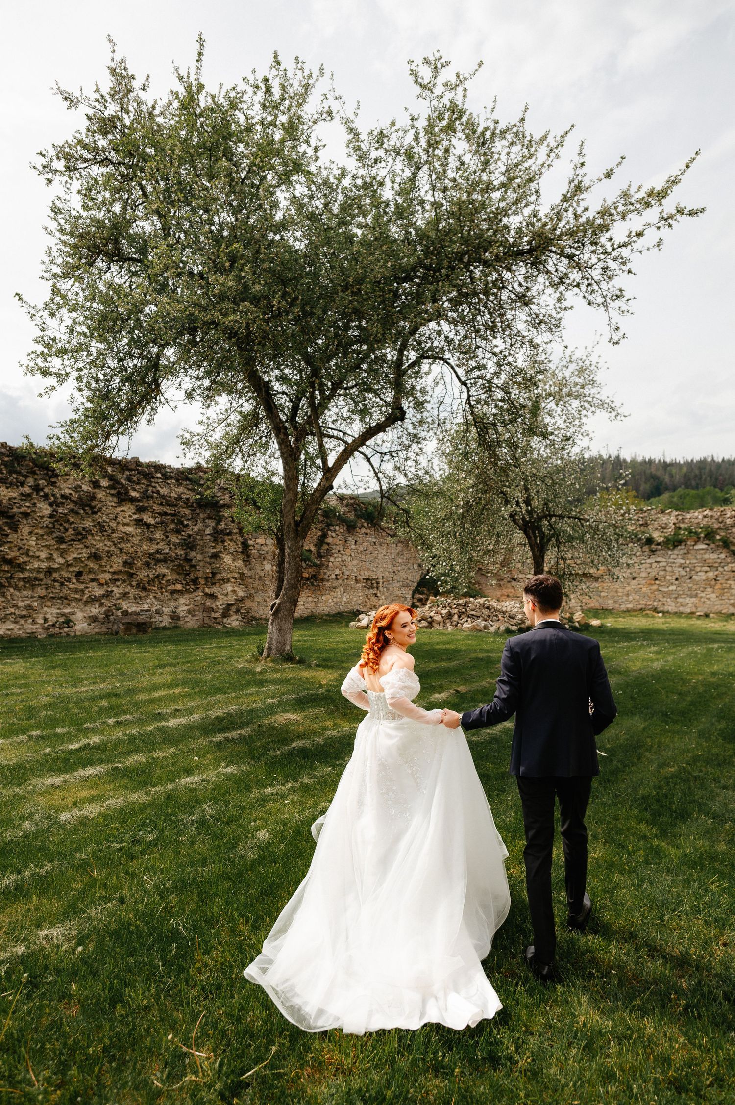 Bride in white gown and groom in black suit hold hands walking toward blooming tree on green lawn.