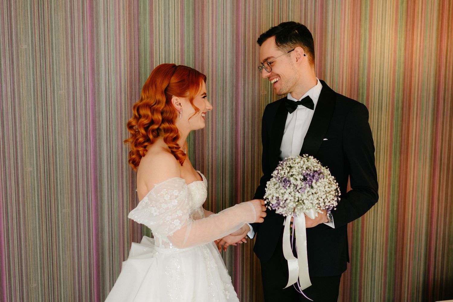 Bride in white gown and groom in black tuxedo exchanging smiles while holding a white bouquet against striped backdrop.