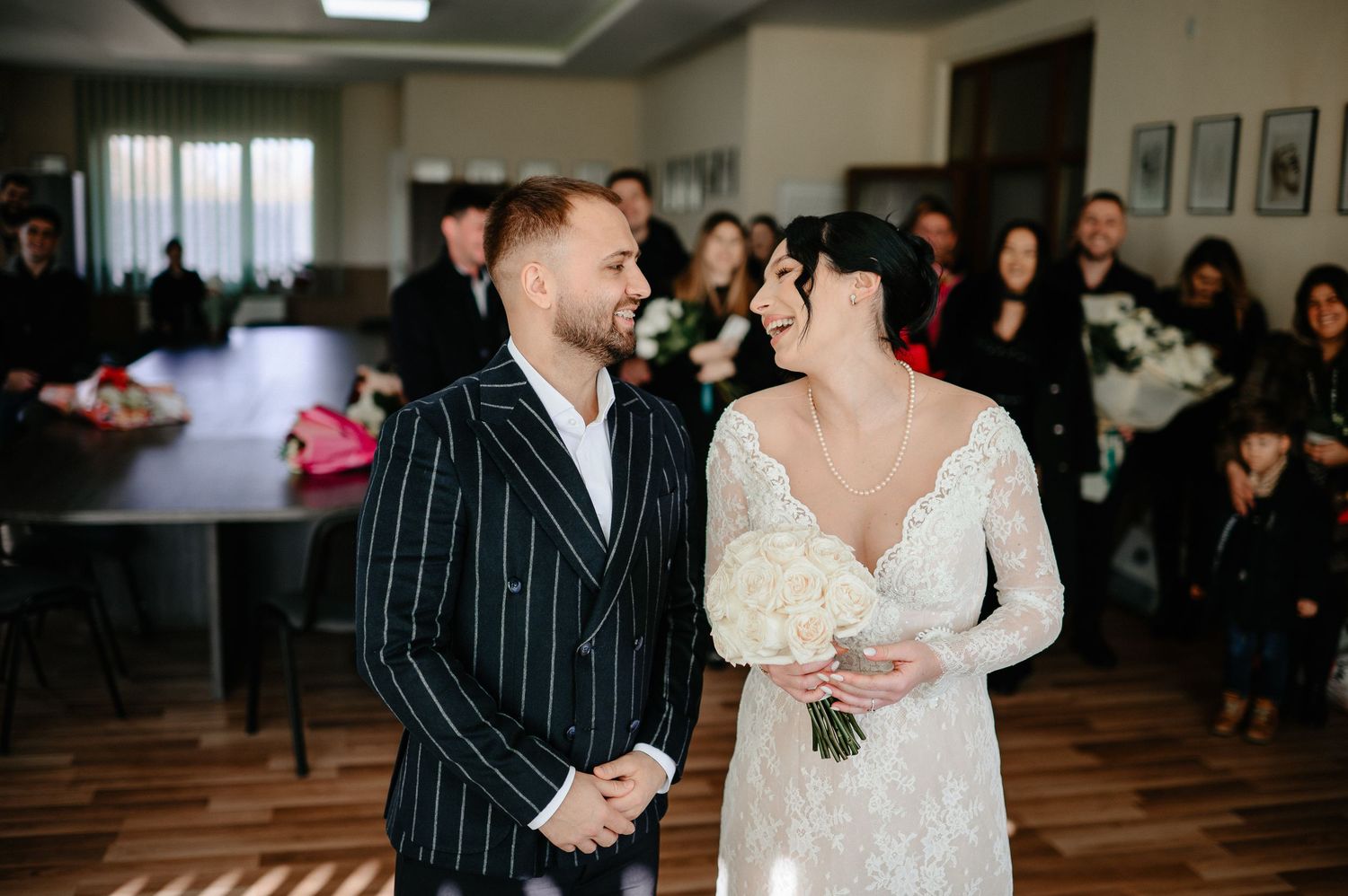 Bride and groom smiling at each other during wedding ceremony, bride in lace gown holding white bouquet.