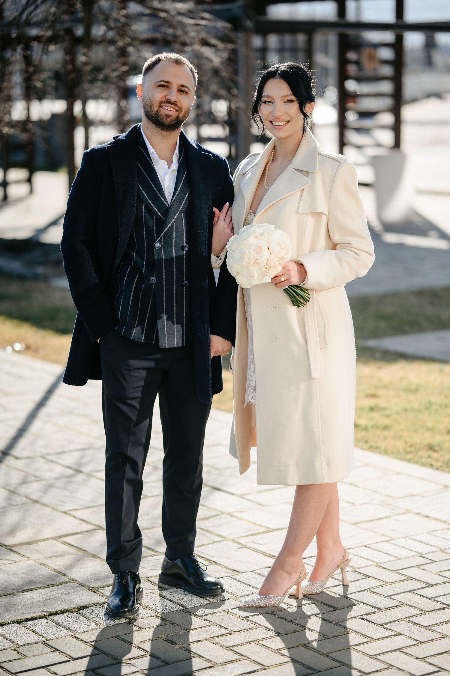 Smiling couple in elegant attire, bride holding white bouquet outdoors.