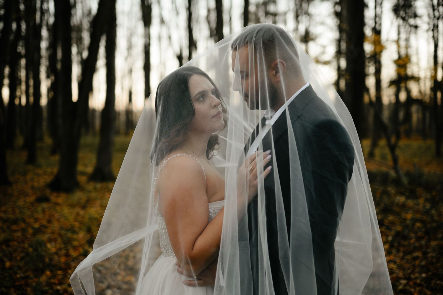 Bride and groom share intimate moment under wedding veil in autumn forest at golden hour.