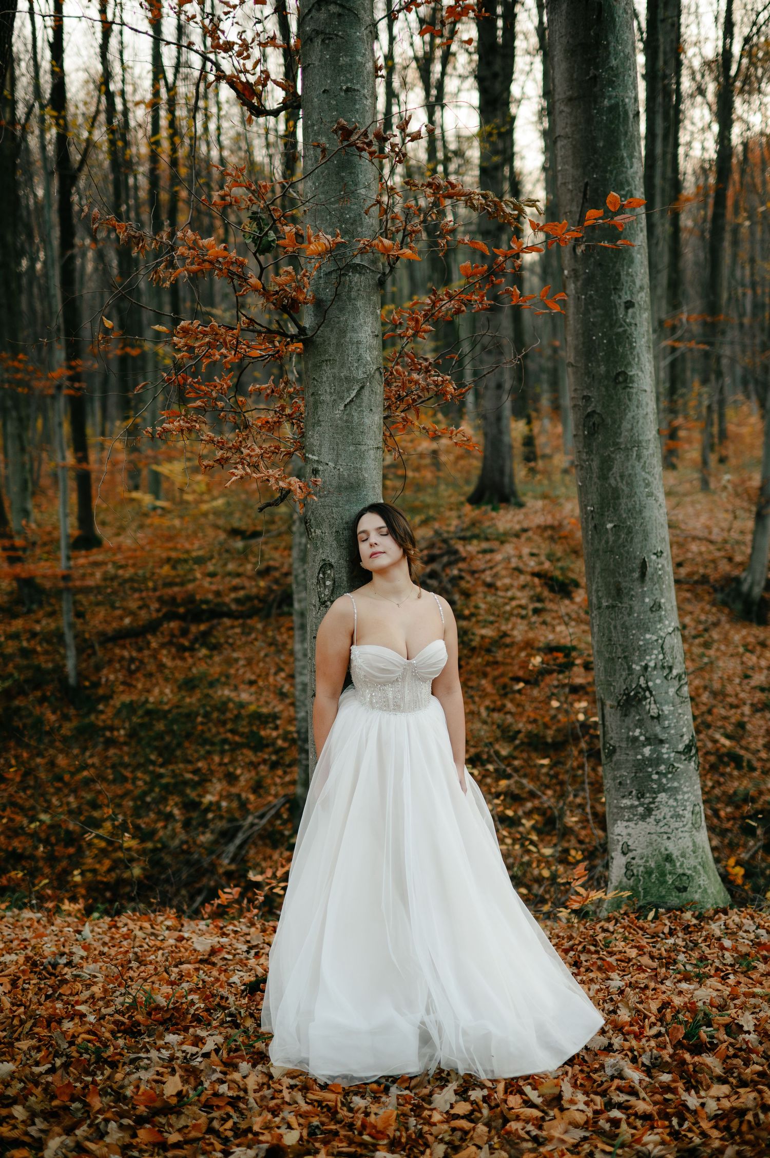 Woman in white strapless wedding gown standing among autumn forest trees with orange fallen leaves.